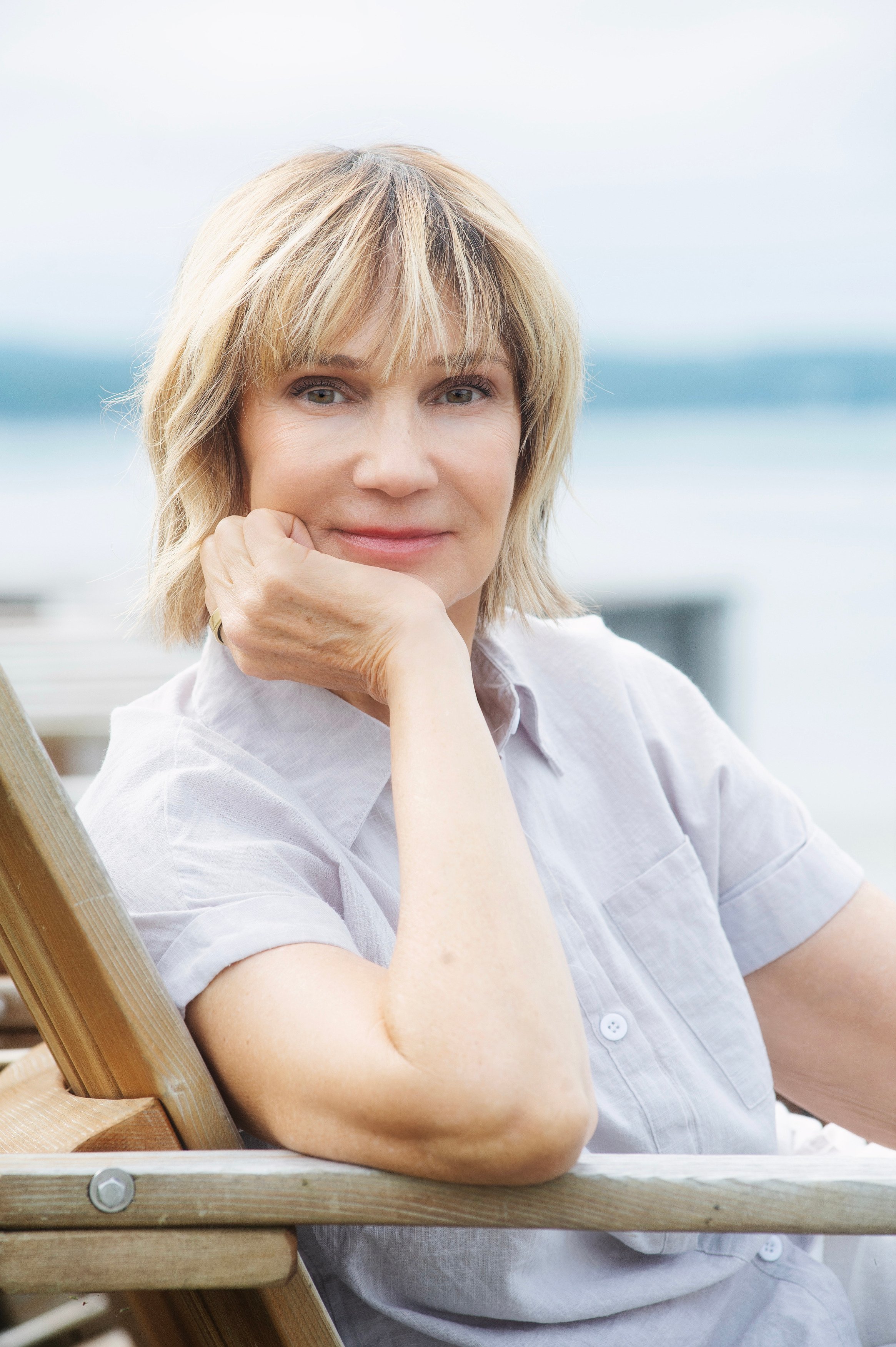 A woman with blonde hair resting her chin on her hand, sitting outdoors near the water.