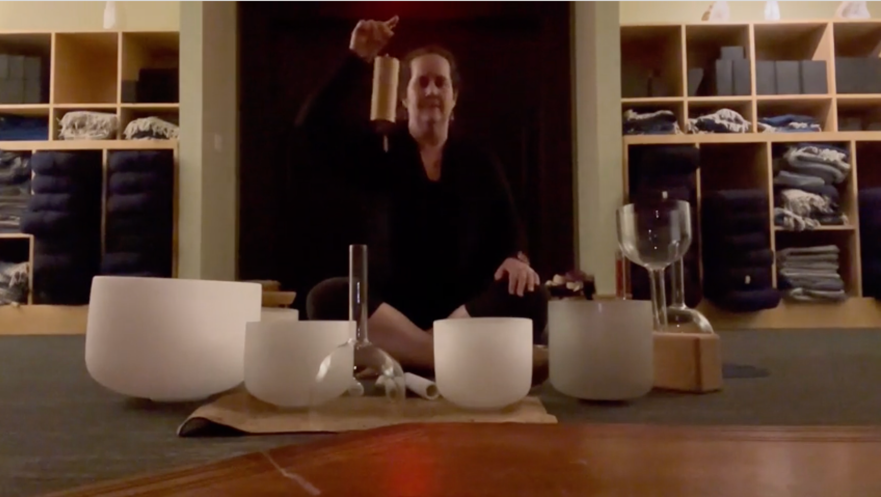 Person sitting on a table behind glass singing or speaking with a raised hand, surrounded by singing bowls and glass objects, in a room with wooden shelves of folded towels or clothing.