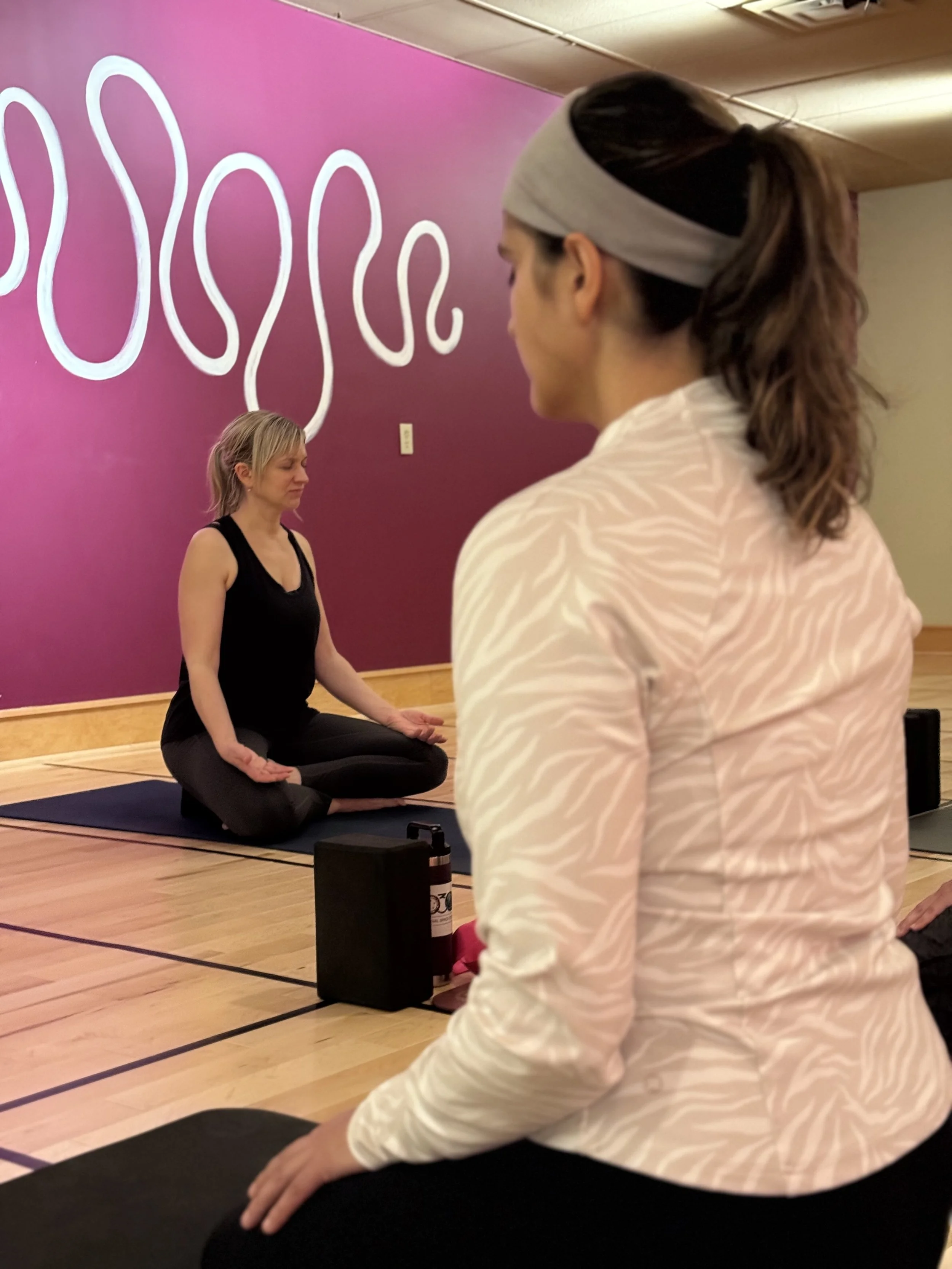 A woman in a black tank top and black leggings is sitting cross-legged on a yoga mat in a meditation pose, eyes closed, in a yoga or fitness studio with a purple wall and abstract white art in the background. Another woman in a white, tiger-striped top and a gray headband, also sitting cross-legged, is observing or meditating.