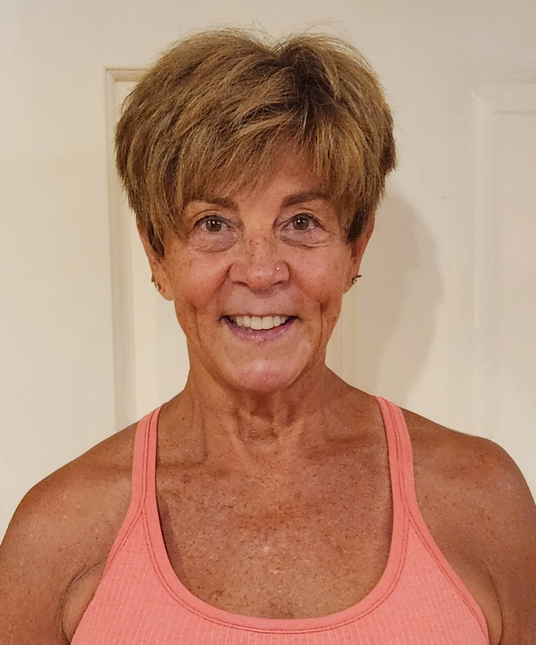 A smiling older woman with short, light brown hair, wearing a pink tank top, standing in front of a white door.