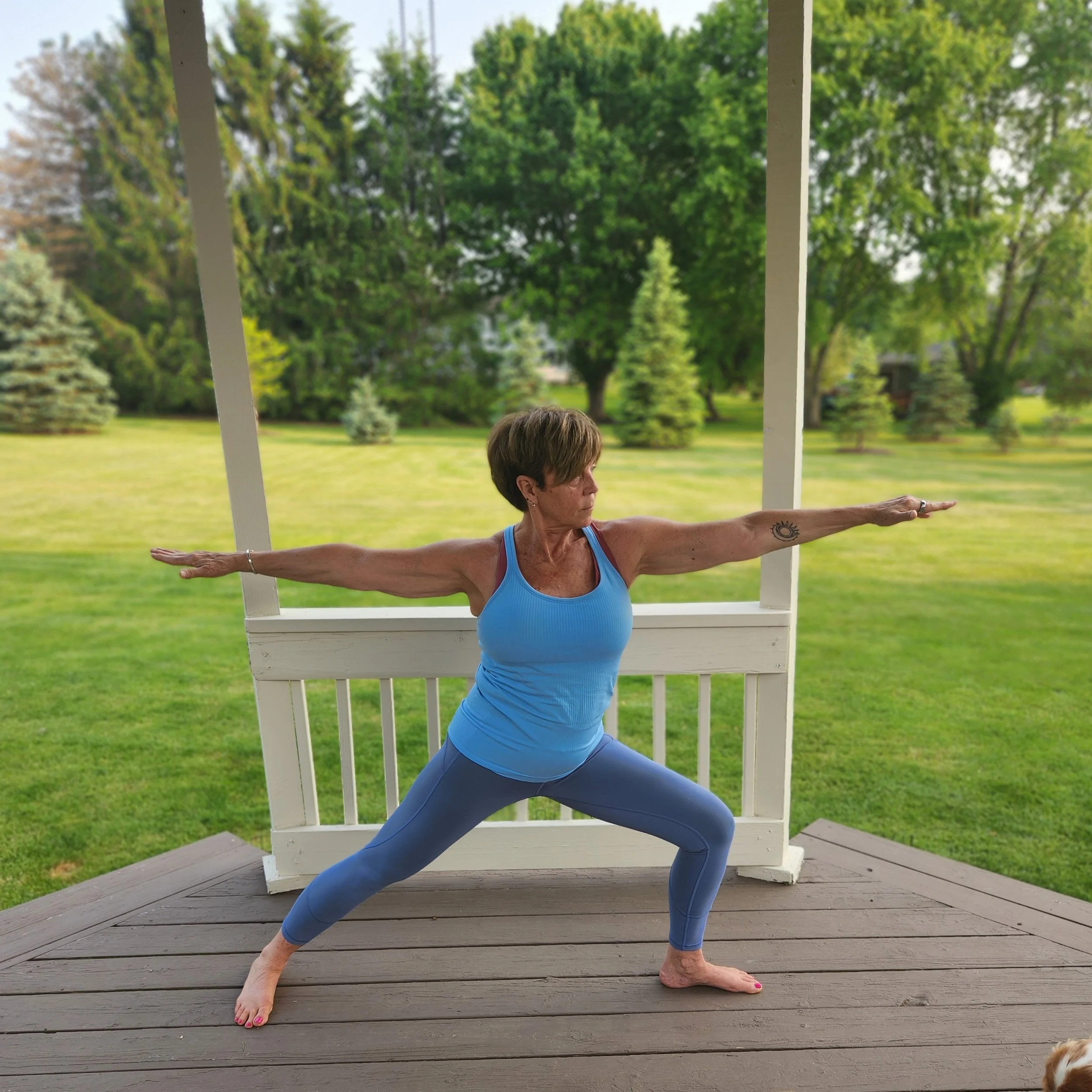 Woman practicing yoga on a porch, standing in Warrior pose with arms extended and feet apart, wearing a blue tank top and matching leggings, with a background of trees and a lush lawn.