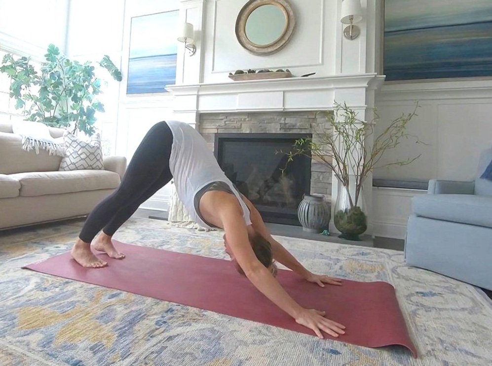 A woman practicing yoga in a living room, performing downward dog pose on a pink yoga mat. The room has a white couch, blue chair, a fireplace, and decorative plants.