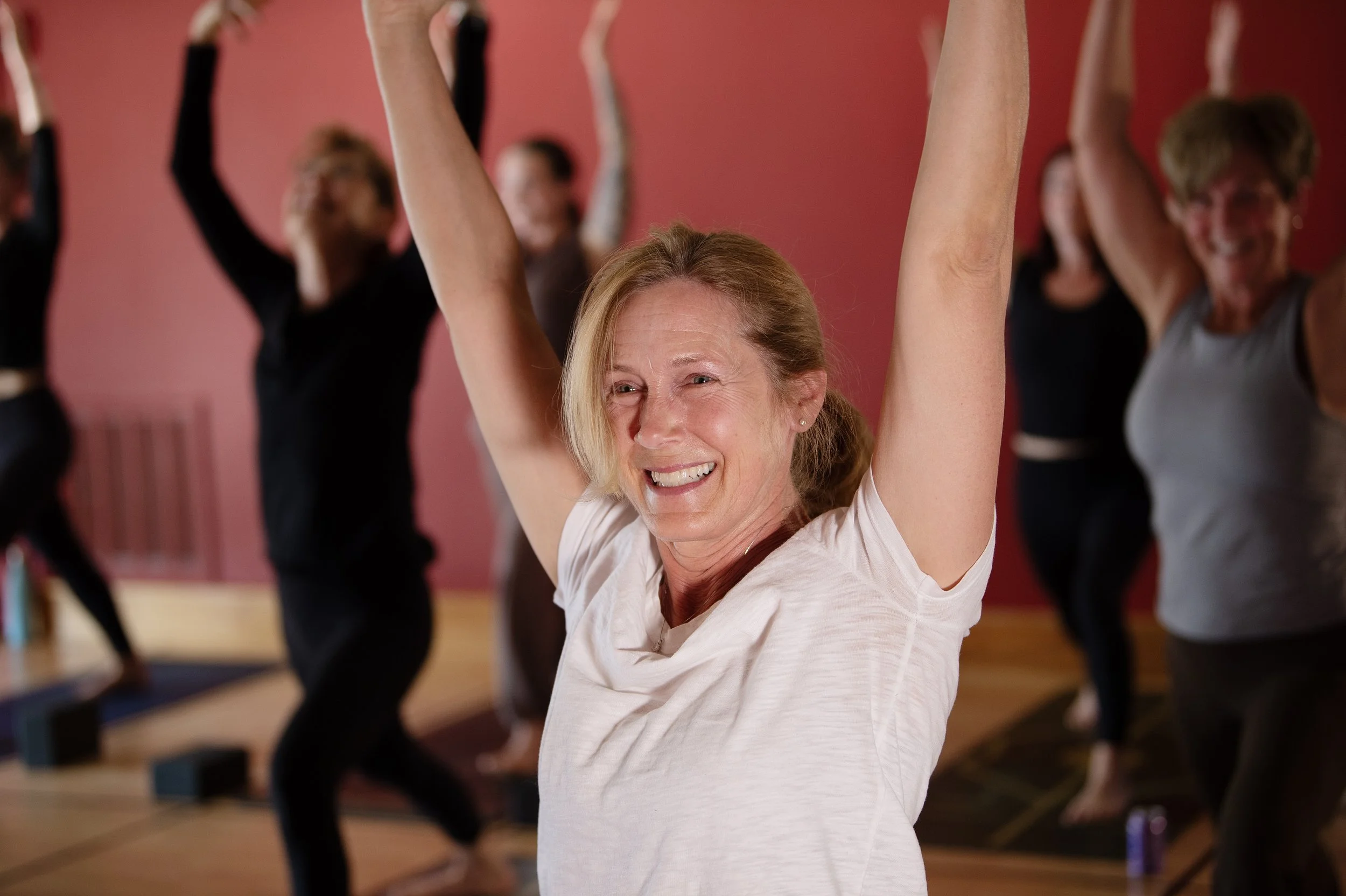 Older woman with red hair smiling while doing yoga with arms raised in a yoga class, group of people practicing yoga in the background.