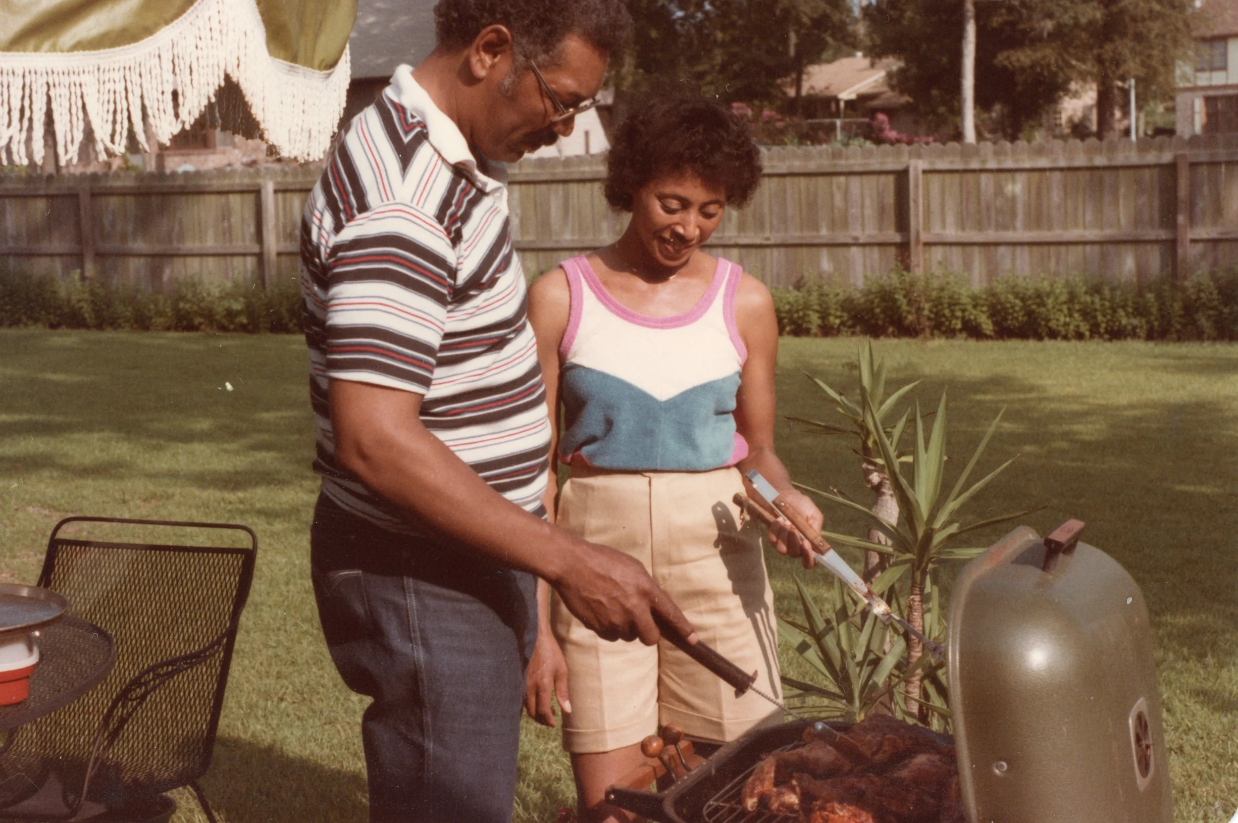 A man and a woman are standing outdoors near a grill, preparing food. The man is wearing a striped shirt and glasses, while the woman is wearing a colorful tank top and beige shorts. There is a fence, trees, and houses in the background.