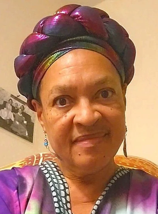 head shot of Black woman in colorful headdress, black and white pic of ancestors on wall in background