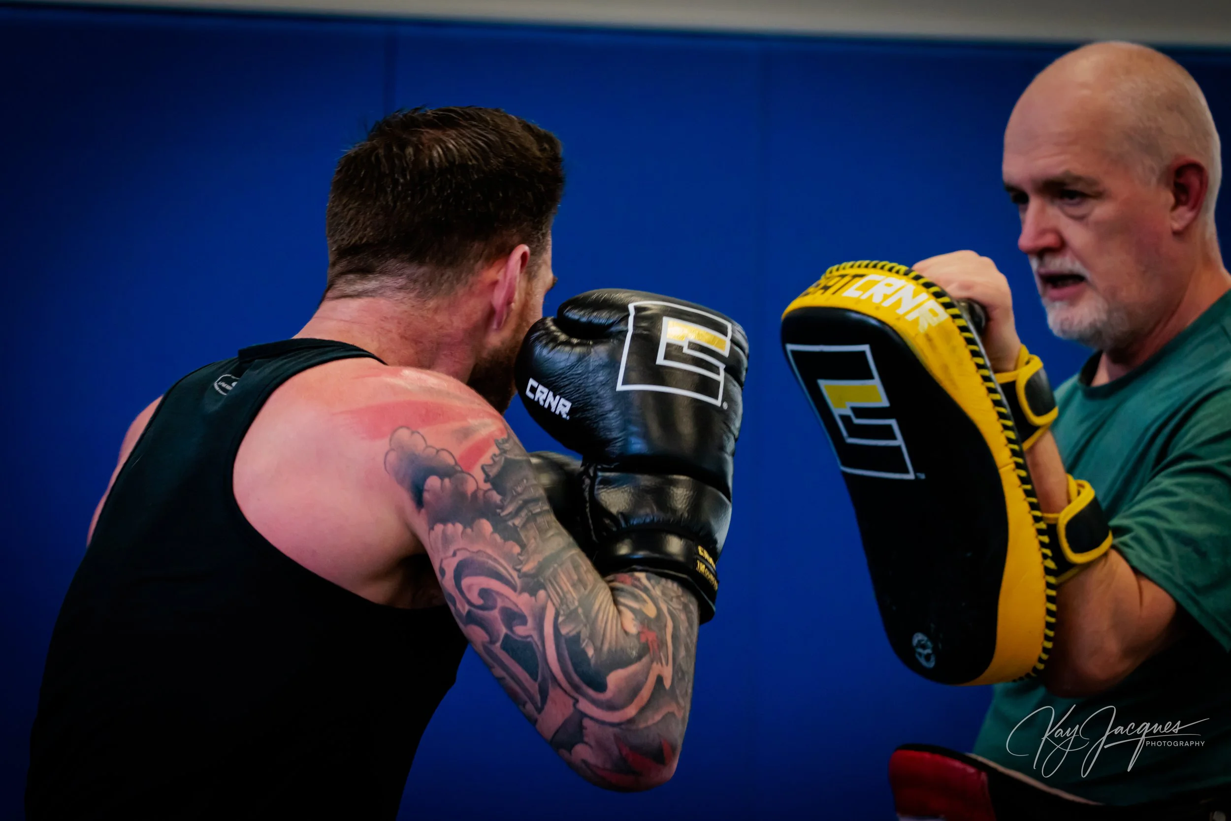 A boxer in a sleeveless shirt with tattoos on his arm practices boxing with a trainer holding focus mitts. The background is blue.