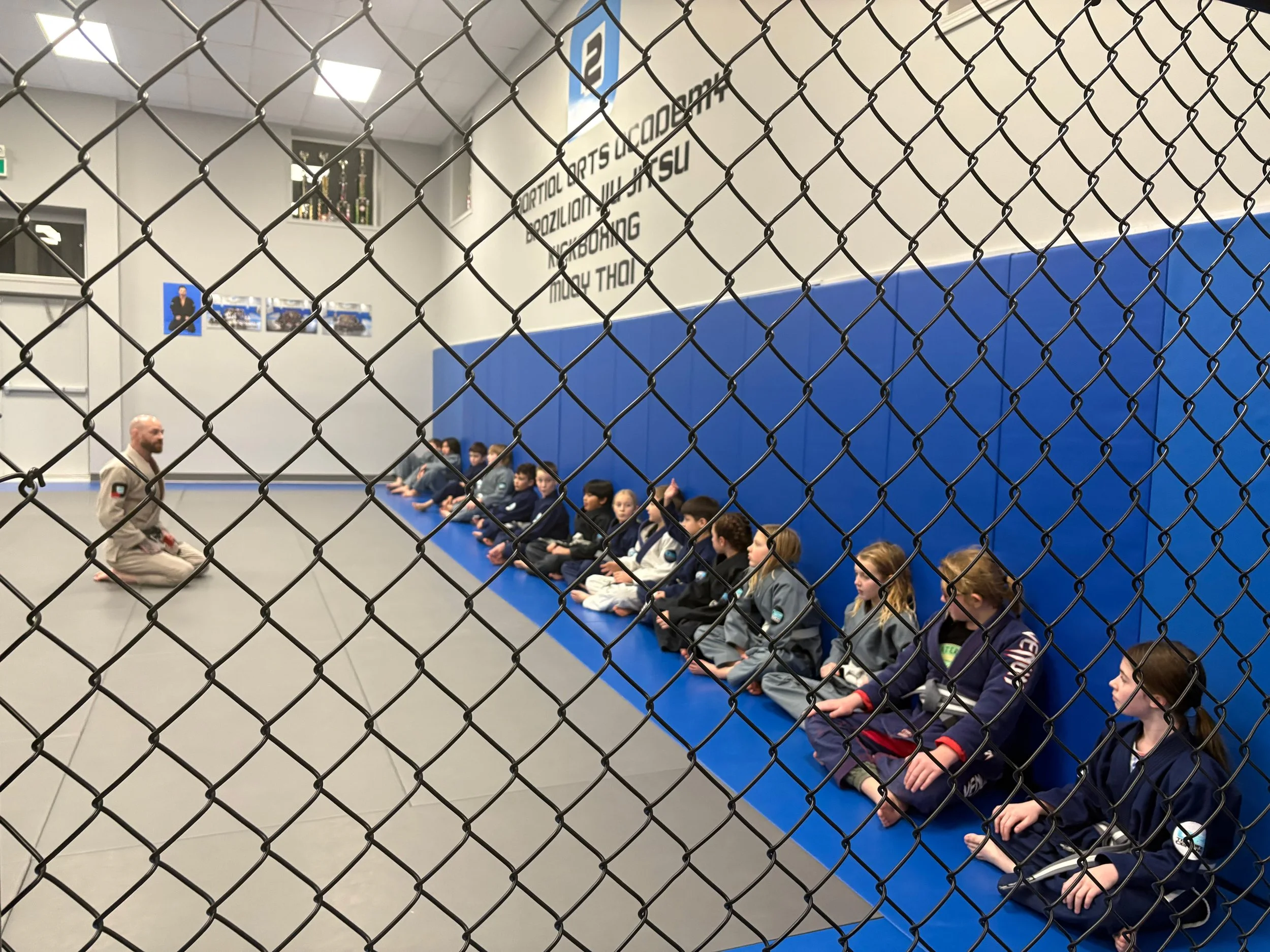 Martial arts class inside a gym viewed through a chain-link fence, with students sitting in a row on blue mats and an instructor kneeling in front of them. The background has posters, trophies, and a large sign with martial arts words.