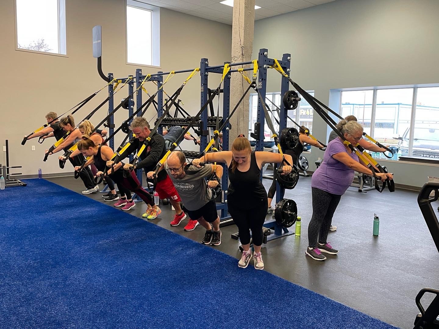 People performing resistance band exercises in a gym, standing in a row and pulling resistance bands attached to an overhead structure.