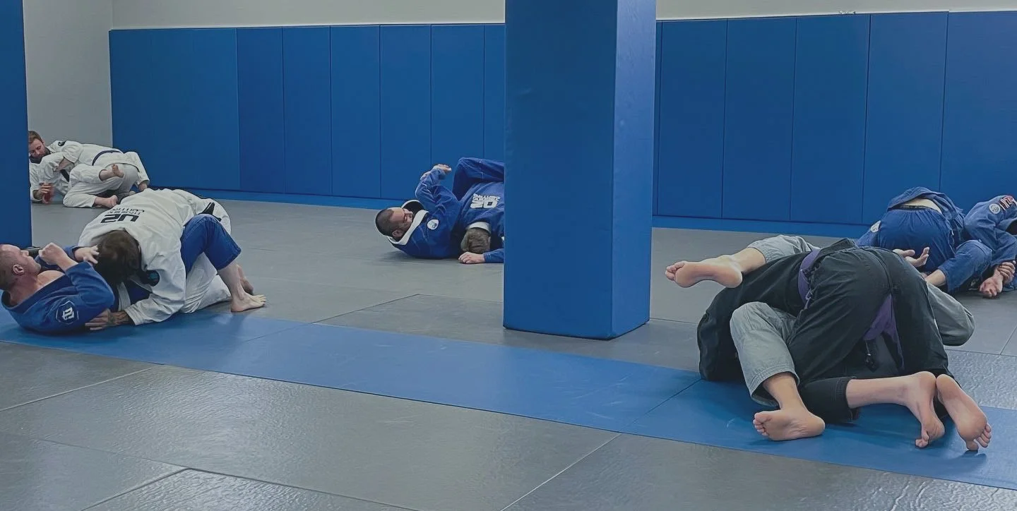 Martial arts students practicing Brazilian Jiu-Jitsu, lying on mats and engaging in grappling drills in a training facility.