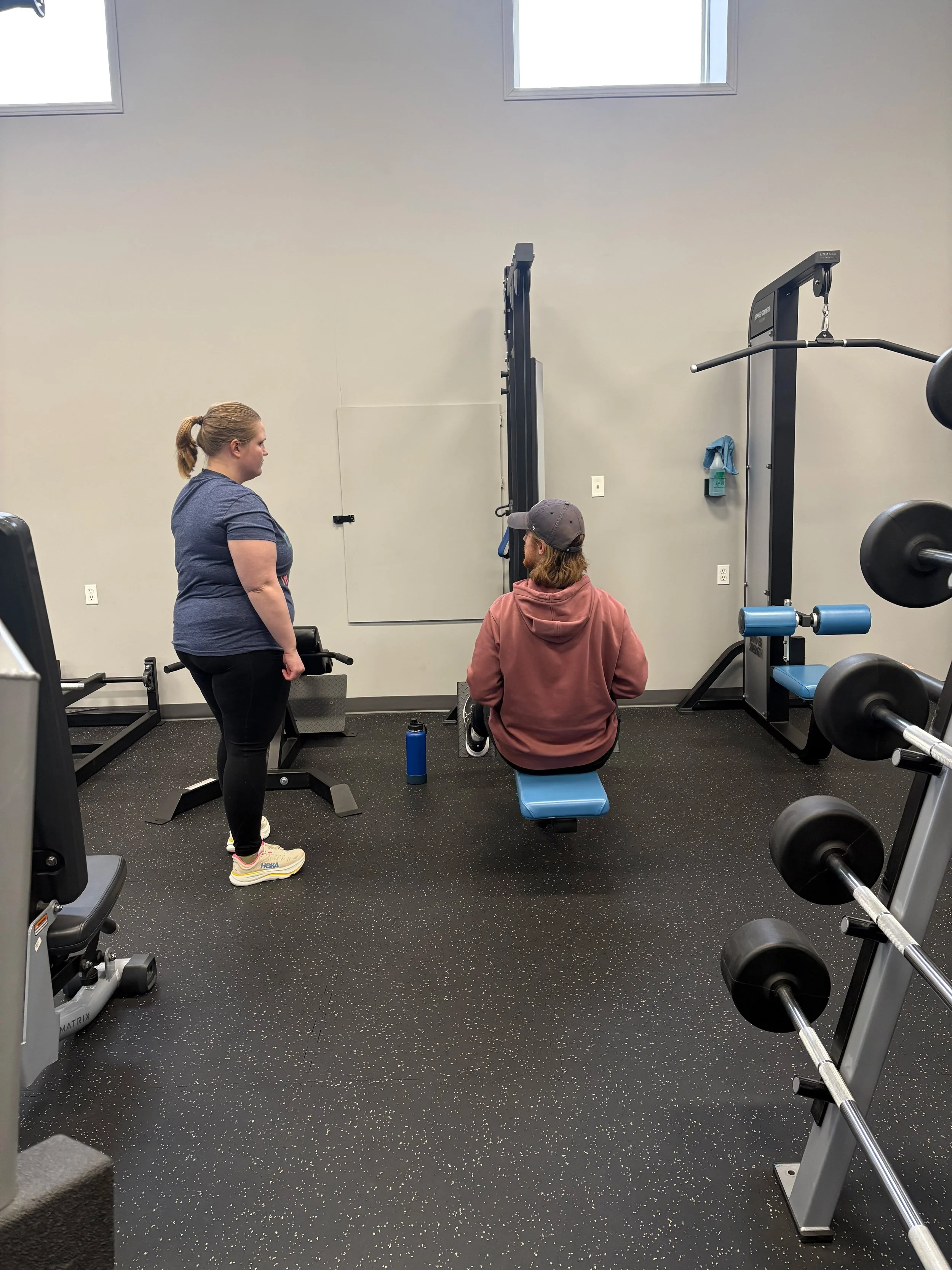 A woman and a man in a gym, with the woman standing and the man sitting on a bench. Various exercise equipment, including barbells and weight machines, are visible around them. The woman appears to be talking to the man, who is looking up at her.
