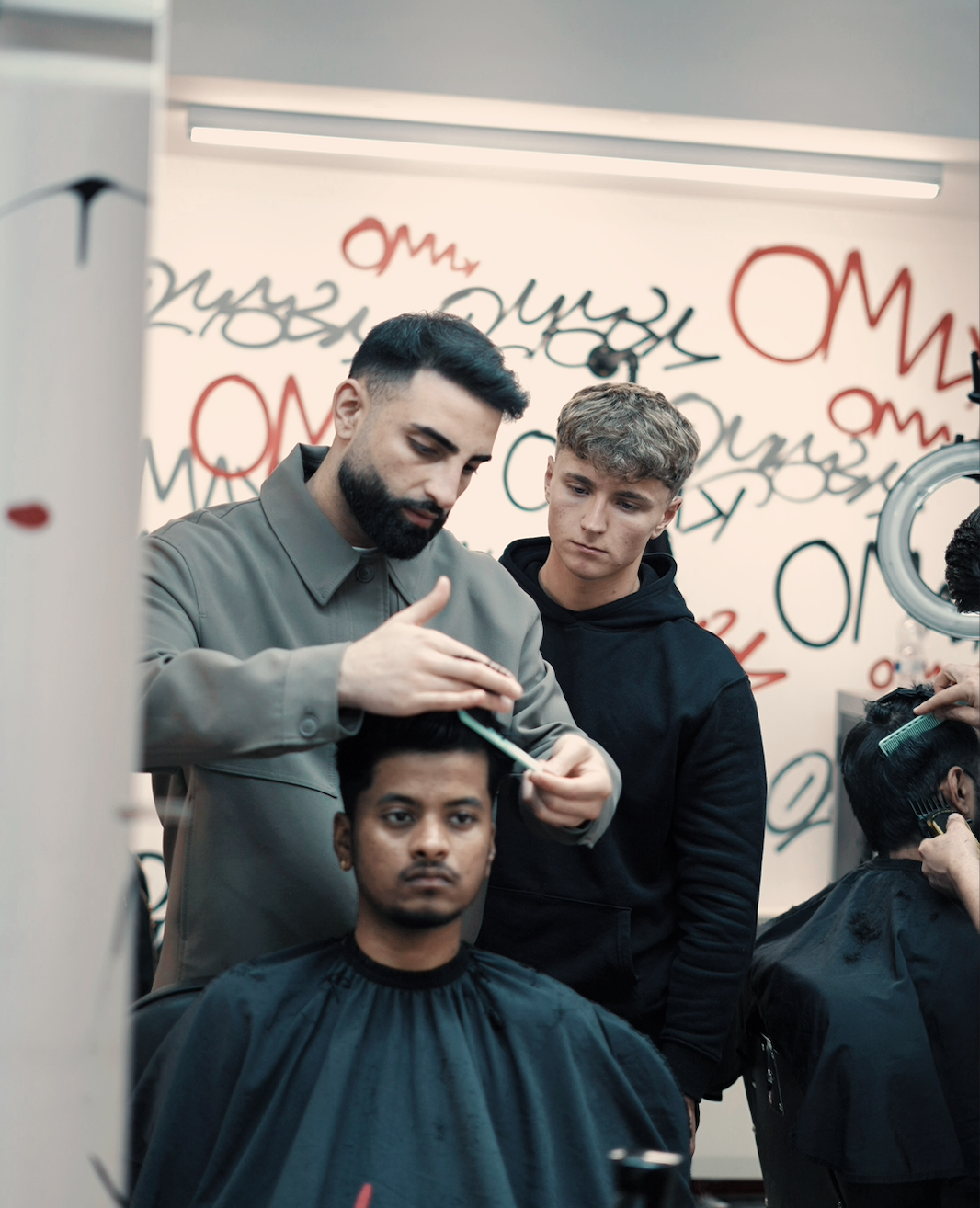 A barber giving a haircut to a male customer while a young man watches, inside a modern salon with graffiti-style artwork on the wall.