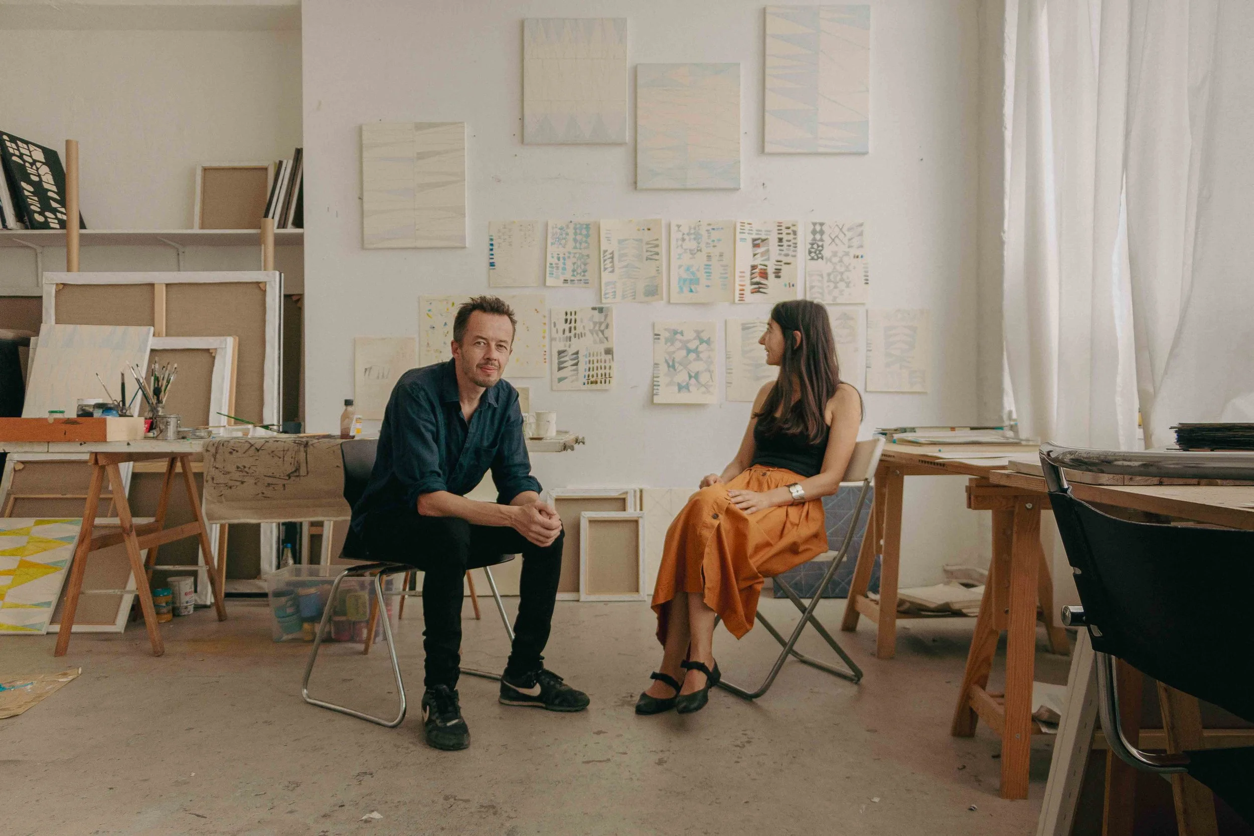 Two people seated in an art studio, surrounded by artworks and art supplies. The man on the left is wearing a dark shirt and black sneakers, while the woman on the right has long dark hair, a black top, and tan skirt, both engaged in conversation.