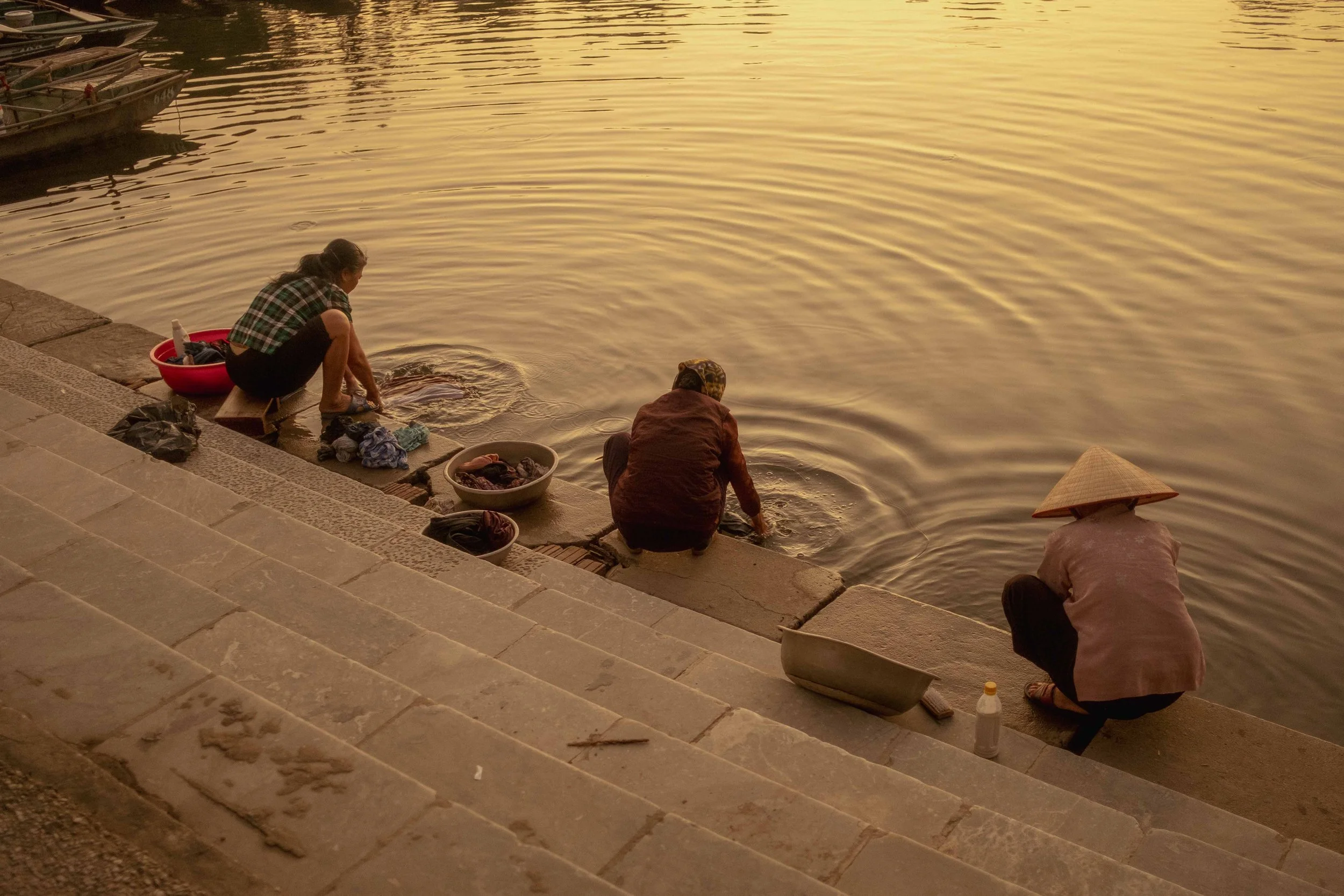 Two people squatting by the river, washing clothes in a bowl at sunset. One person wears a traditional conical hat and the other a checkered shirt, with boats docked in the background.