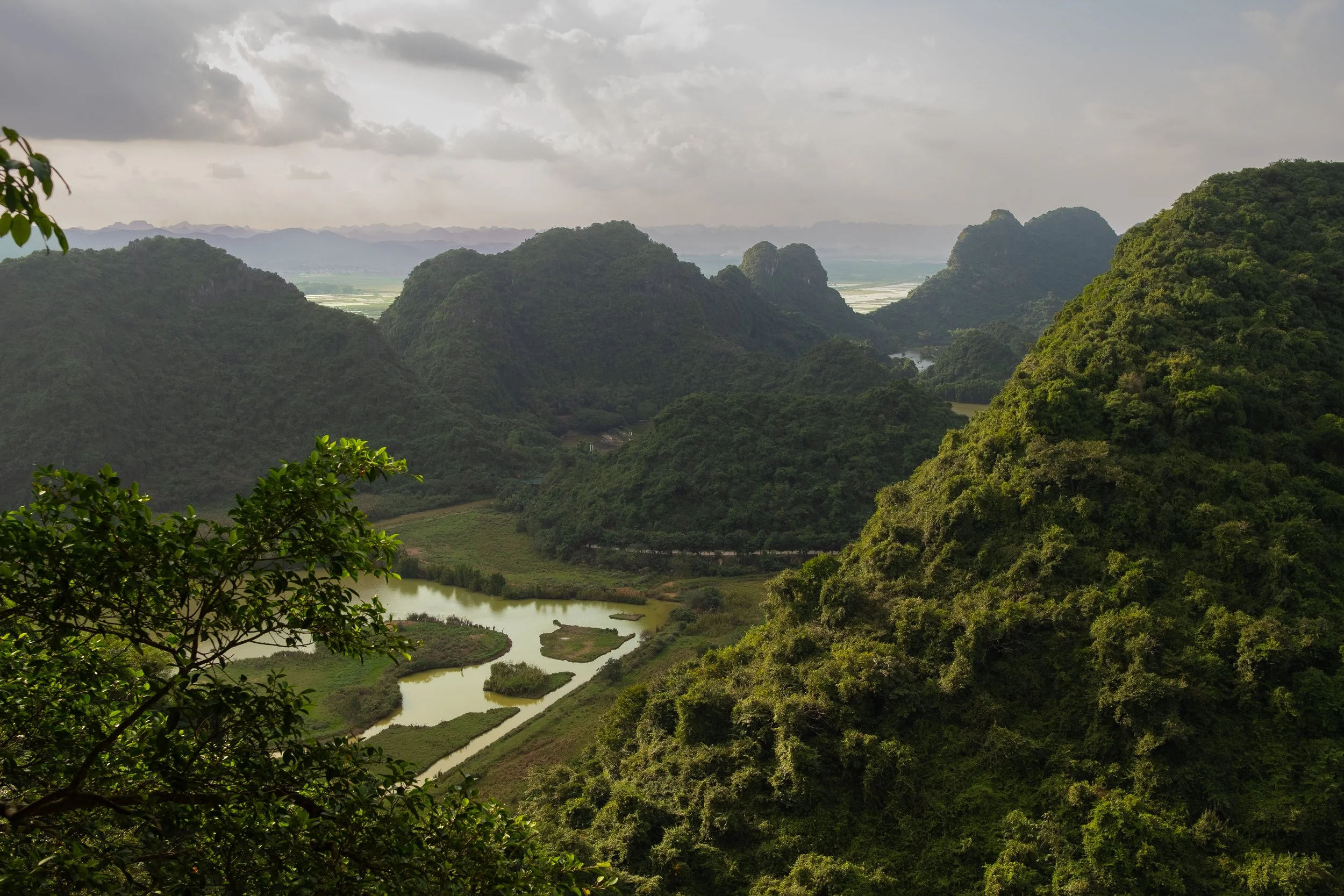 Lush green mountains with a river and farmland below under a cloudy sky.