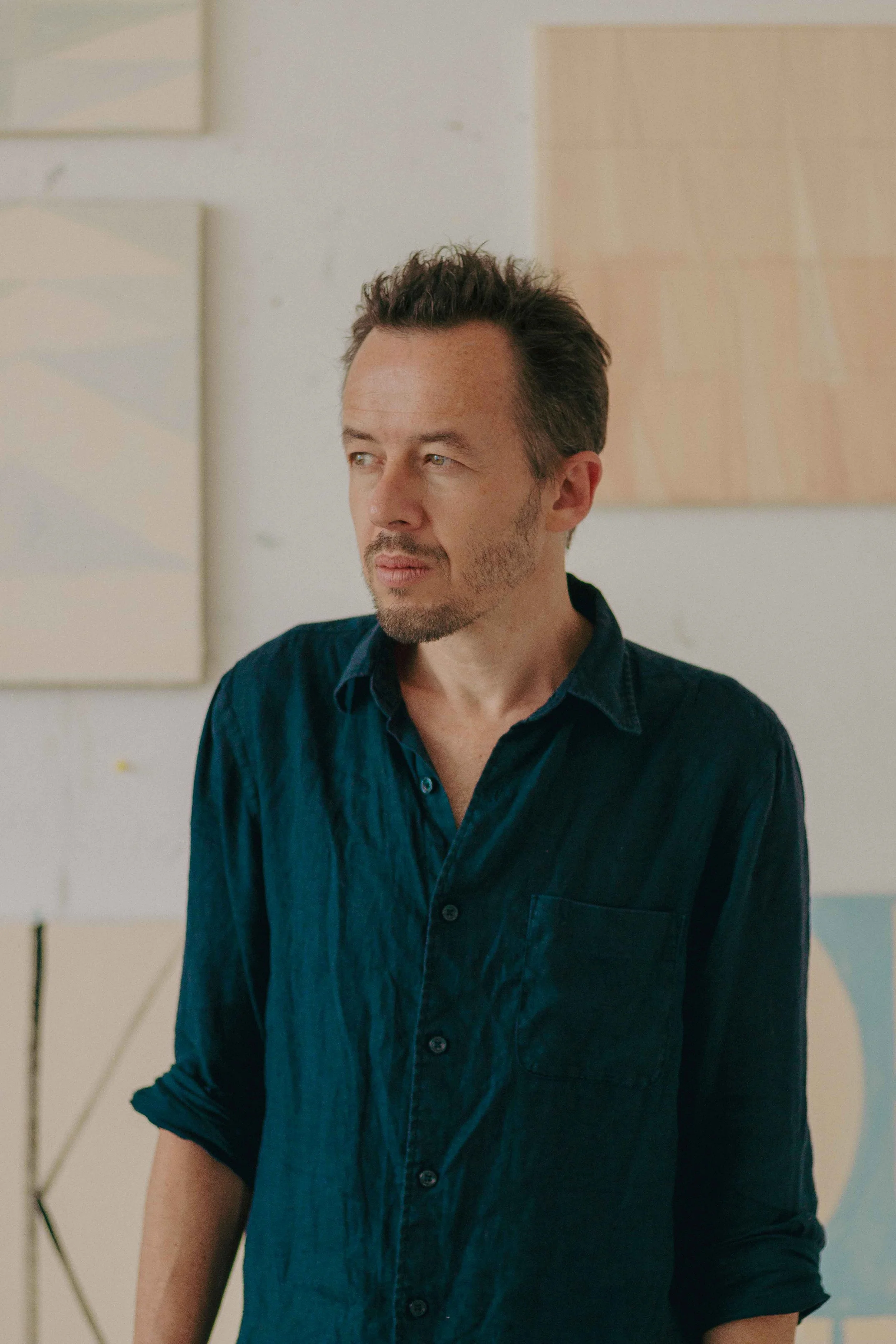 A man with short dark hair and stubble looking to his right, wearing a dark blue shirt with rolled-up sleeves, standing indoors against a background with light-colored abstract art.