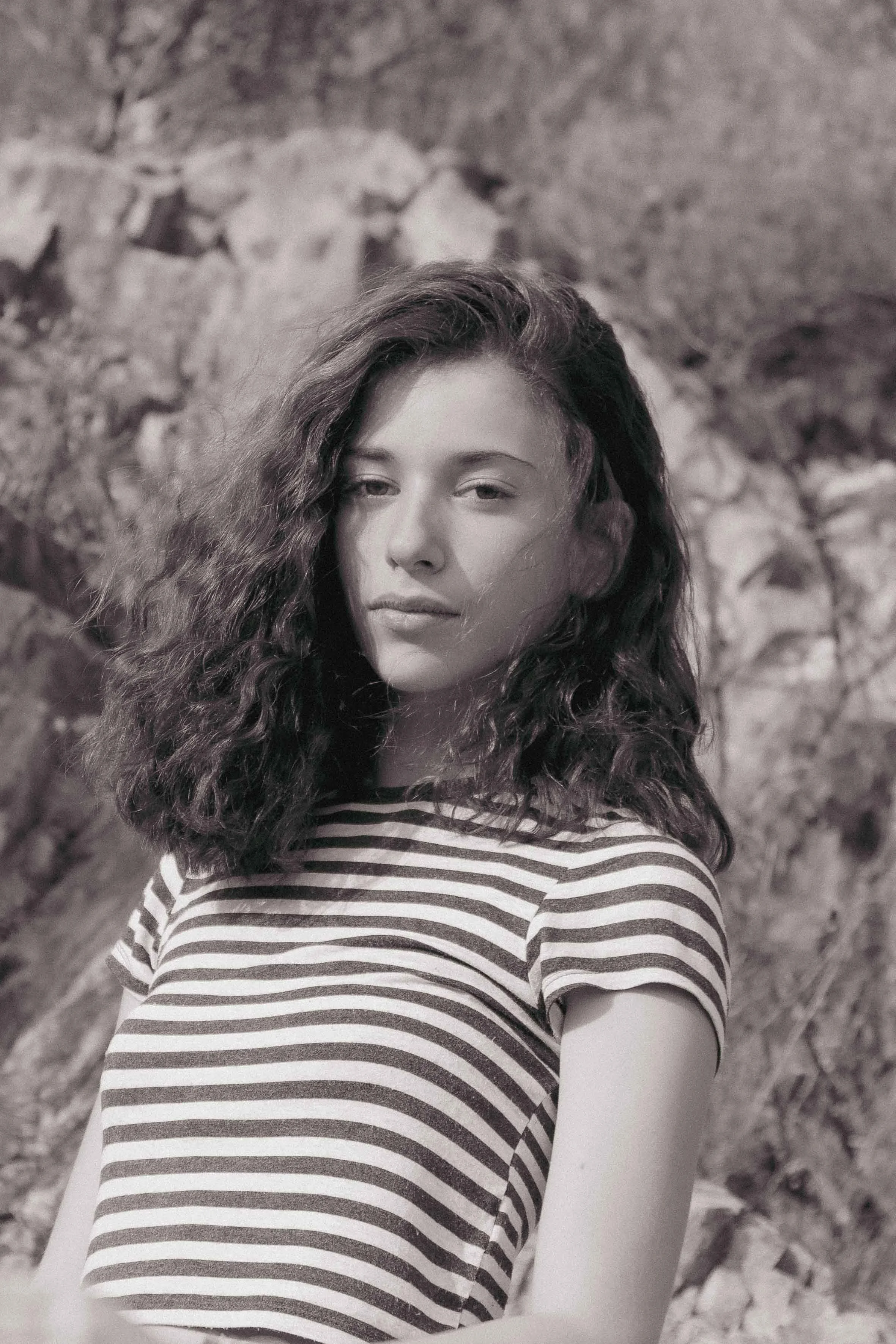 A woman with curly hair taking a selfie outdoors, wearing a striped shirt, with a rocky background.