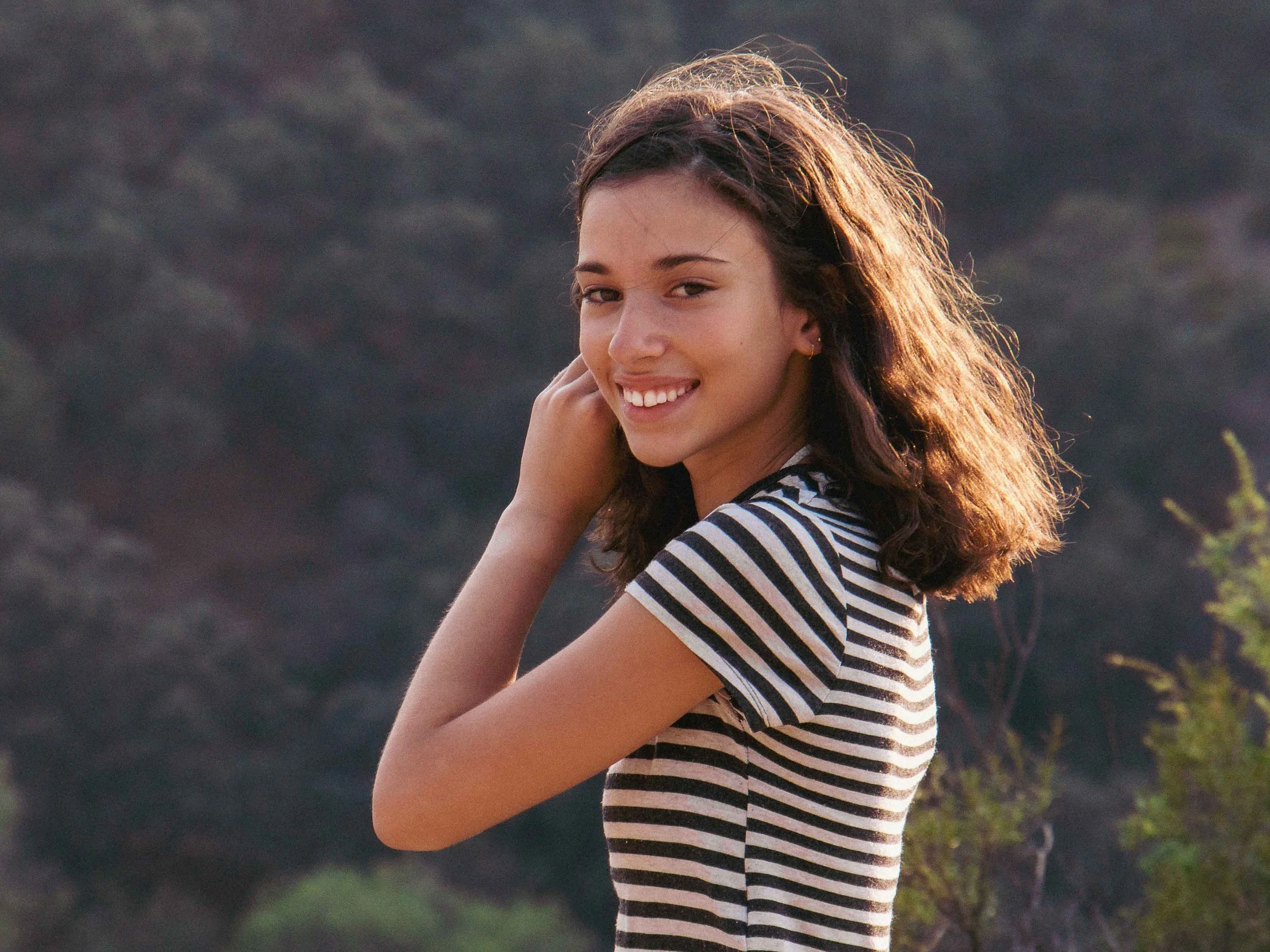 A young woman with long brown hair and fair skin smiling at the camera, wearing a black and white striped shirt outdoors during daytime.