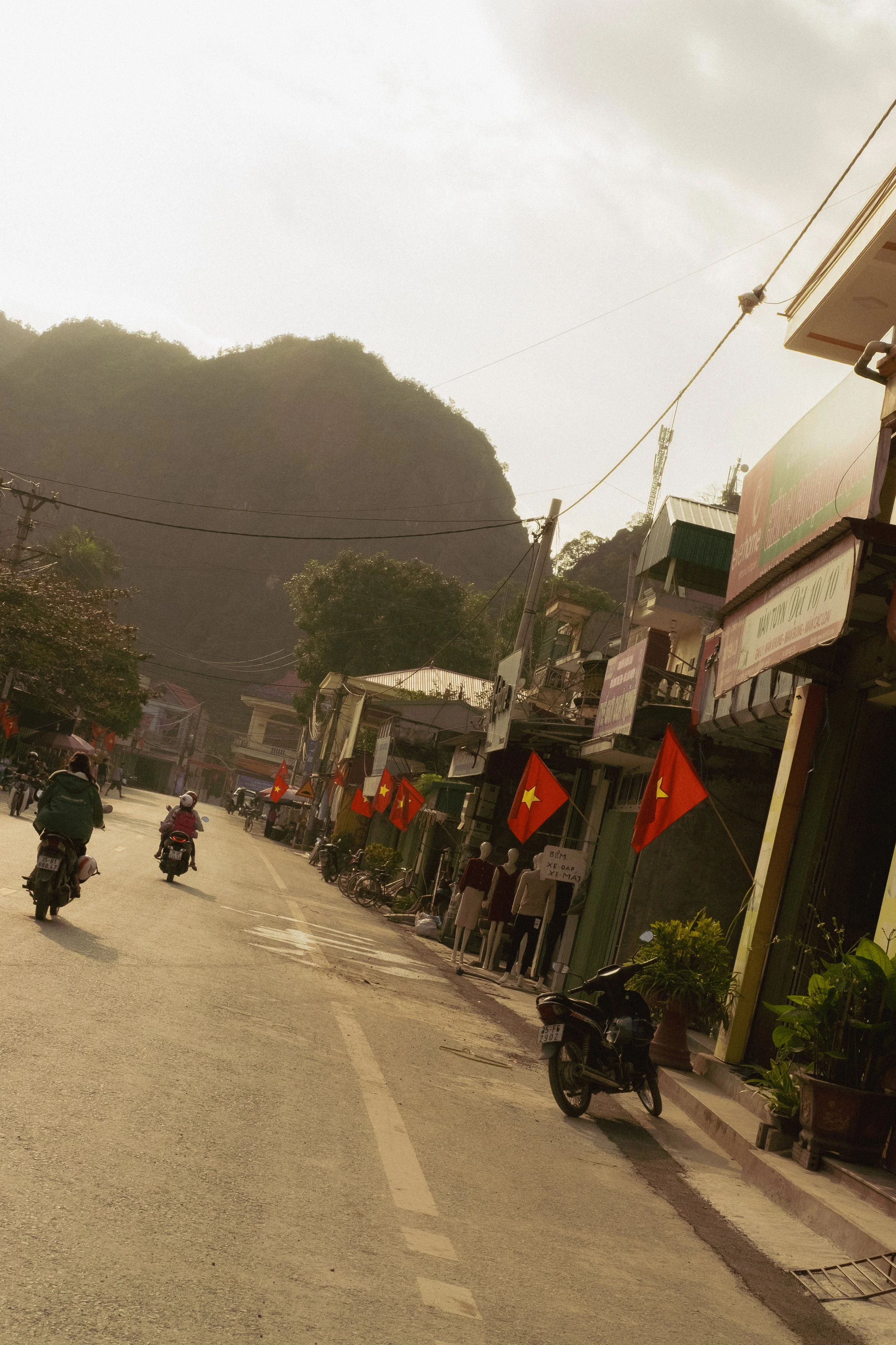 A street scene in a small town with several motorcycles parked and a few people walking. There are flags with yellow stars on red backgrounds hanging along the street. Buildings with storefronts and signs, some with awnings, line the street. In the b