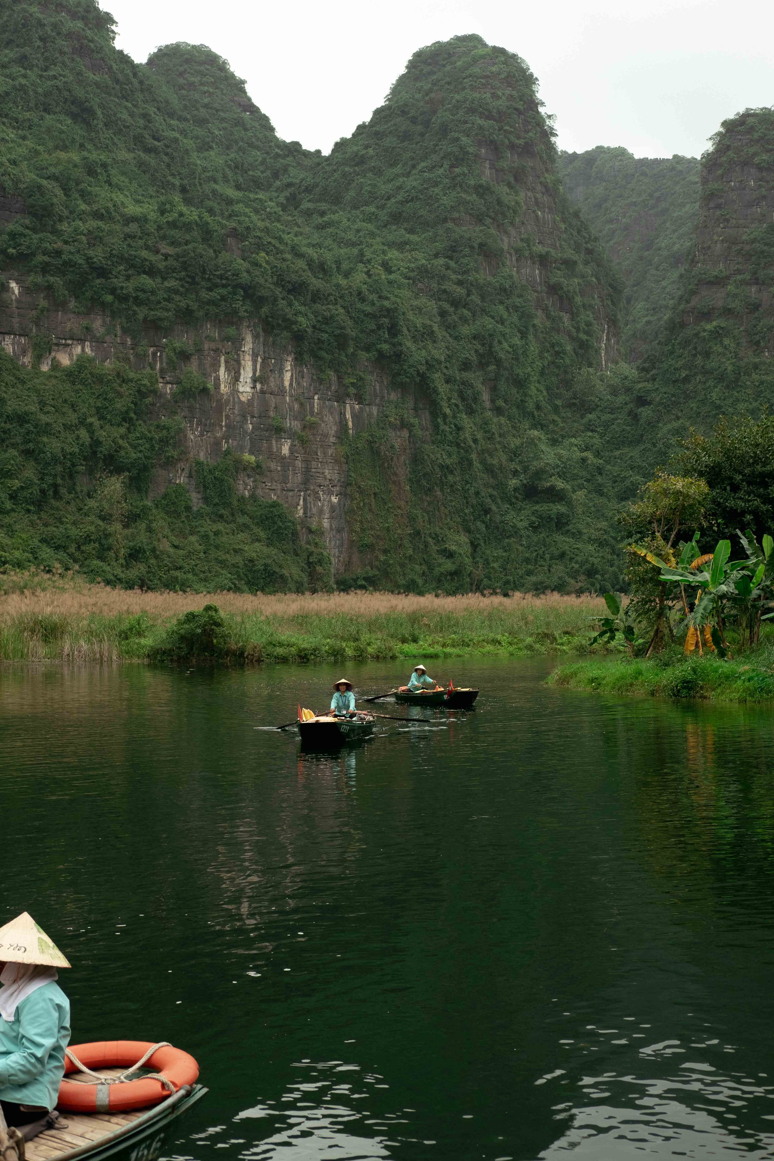 Two people in small boats rowing on a river with lush green cliffs and trees in the background.