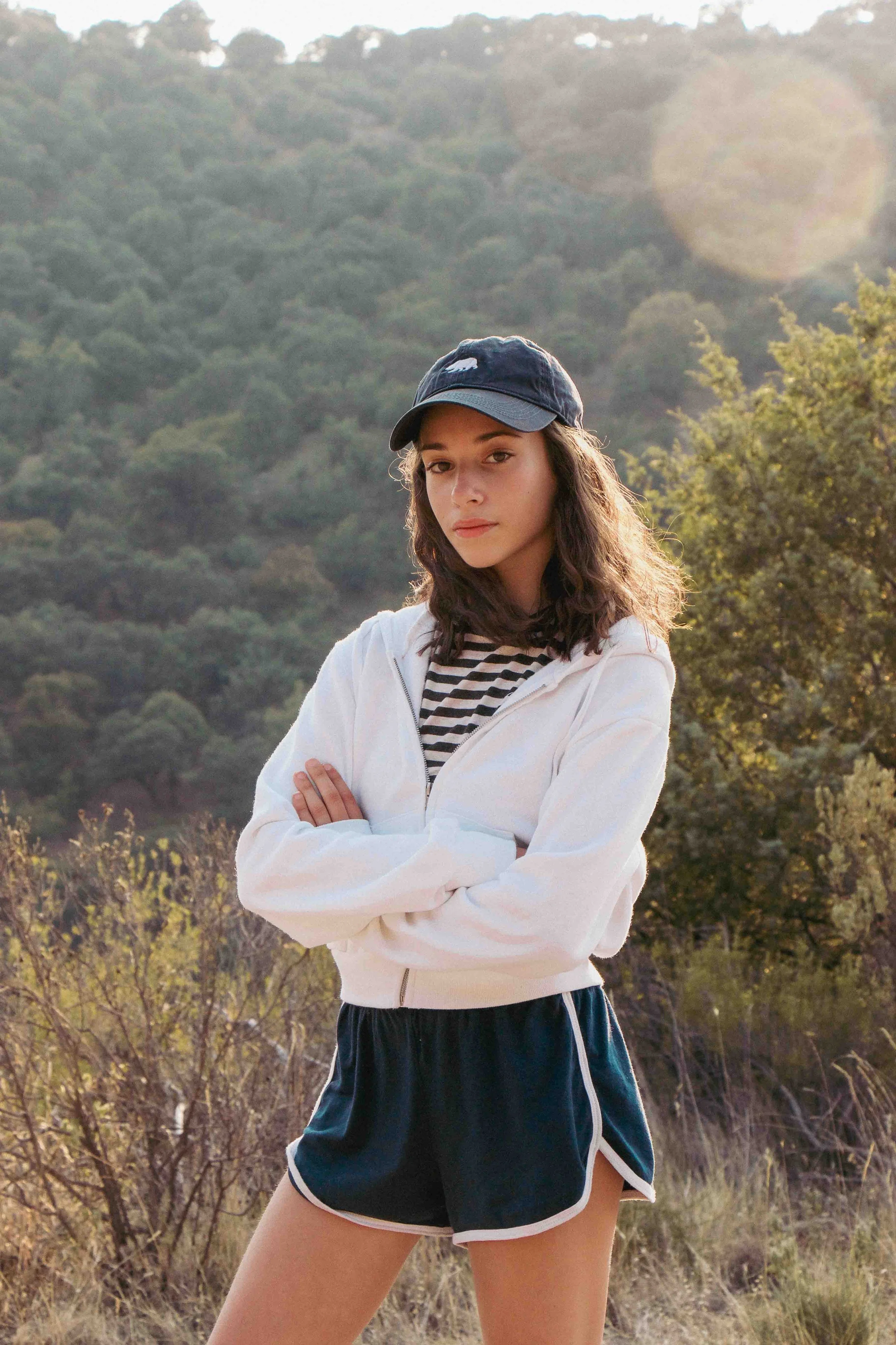 A young woman standing outdoors on a hillside with mountains and trees in the background, wearing a navy cap, white zip-up jacket, and black athletic shorts with white trim.