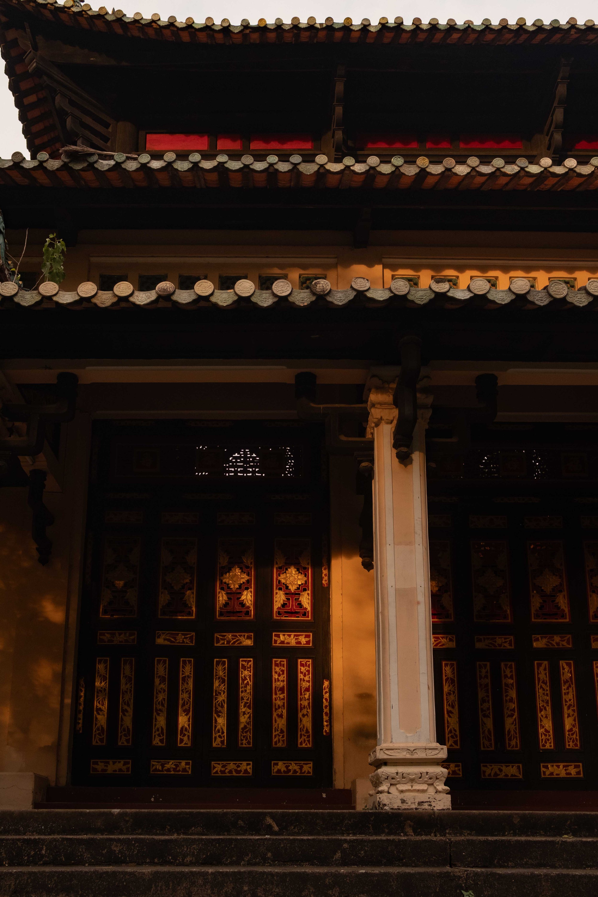 The image depicts the entrance of a traditional Asian temple or building with ornate wooden doors. The doors feature intricate gold designs, and the structure has a pagoda-style roof with layered eaves decorated with small tiles. The warm lighting hi
