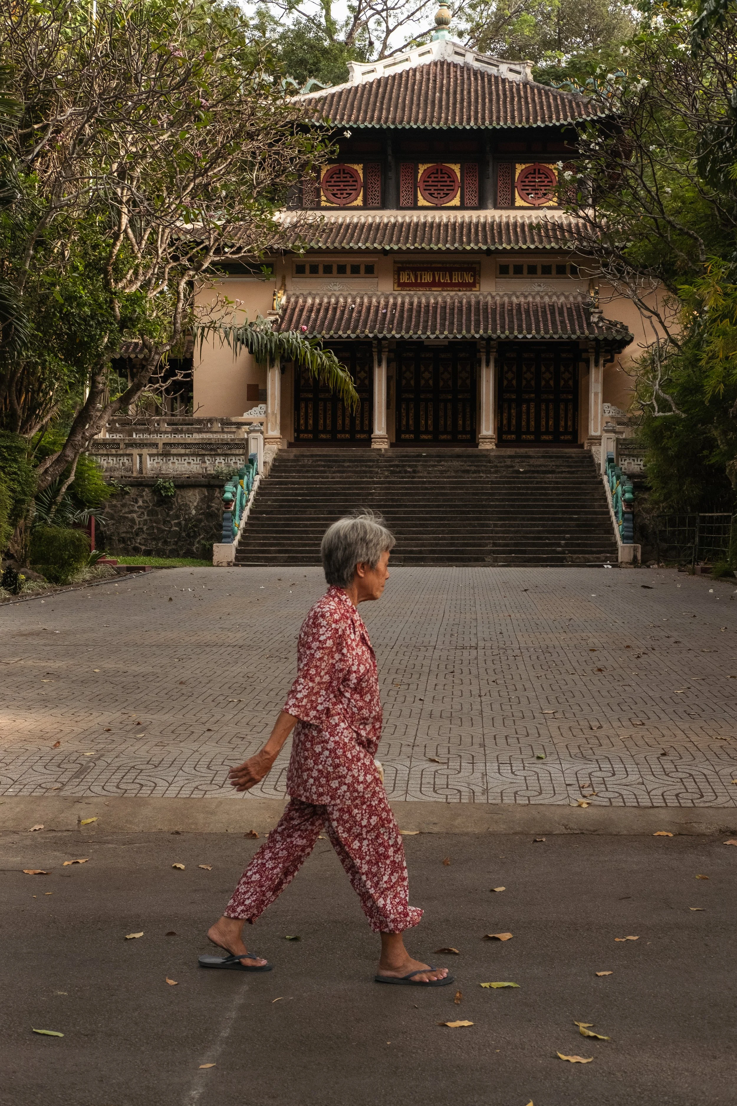 An elderly woman with gray hair, wearing a red floral outfit and black flip-flops, is walking barefoot on an asphalt path with fallen leaves, in front of a traditional Vietnamese temple with a staircase, ornate doors, and decorative circular windows 