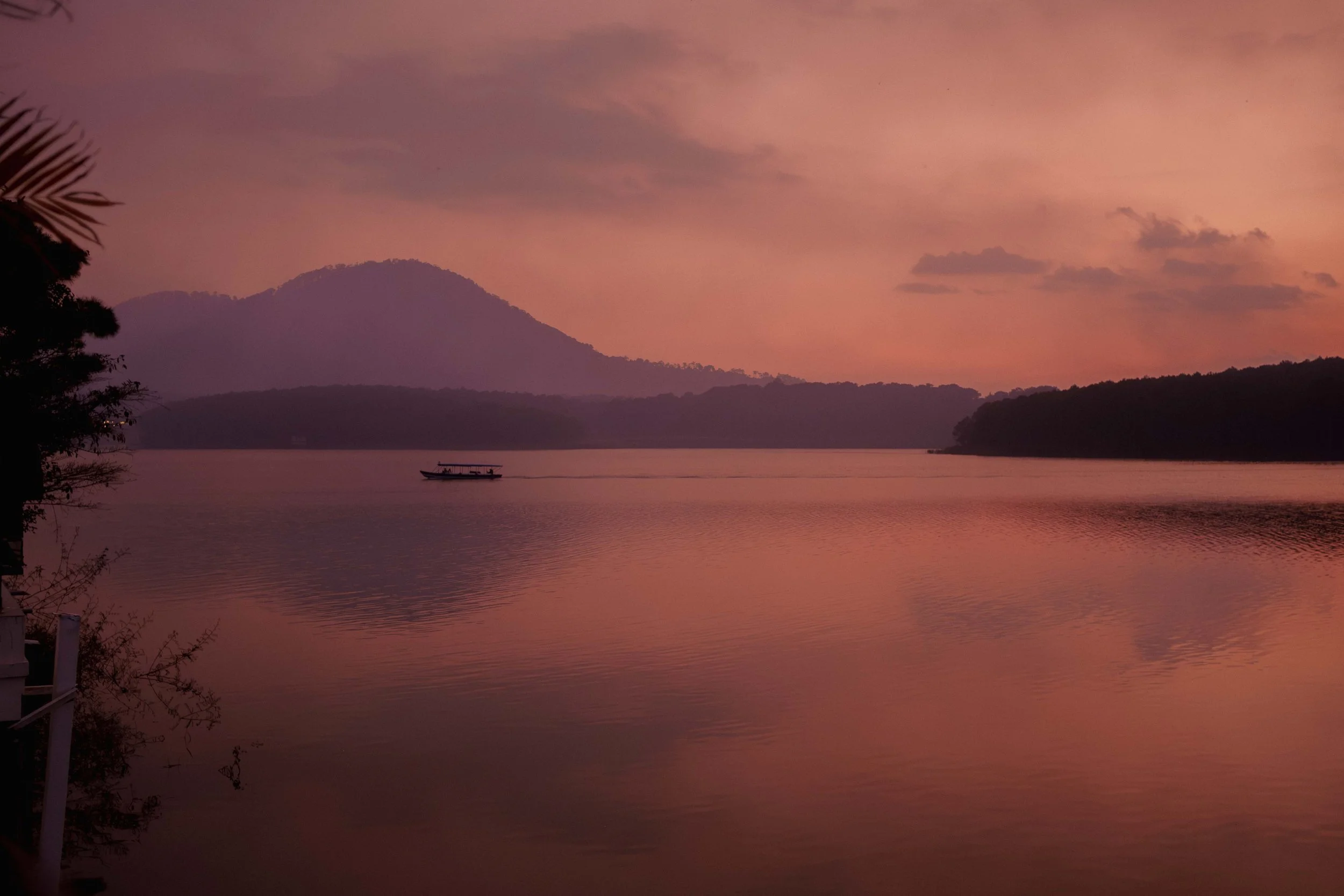 A serene lake surrounded by mountains at dusk with pink and purple sky, a boat on the water, and trees along the shoreline.