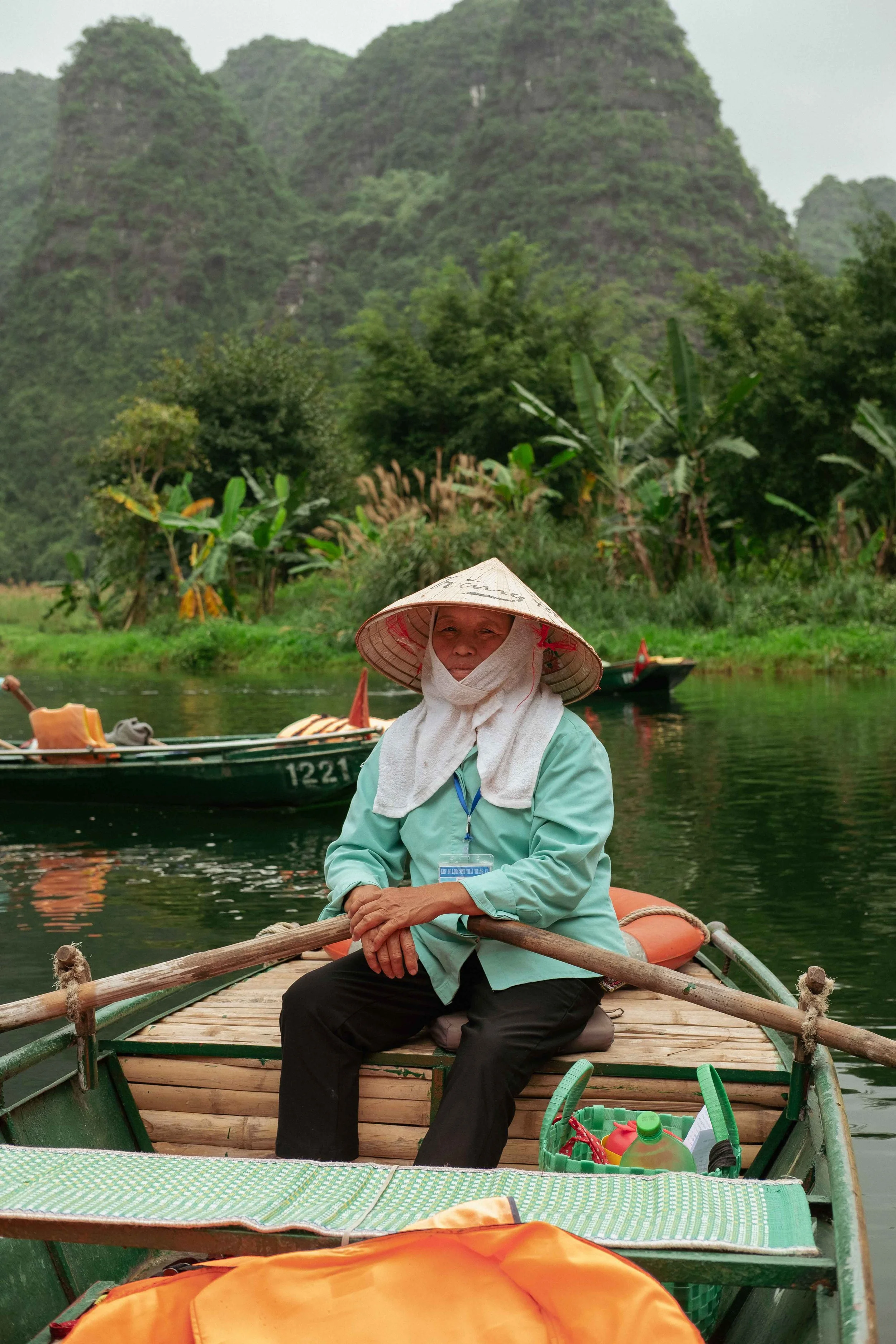 Person sitting on a wooden boat by a river, wearing a traditional conical hat and a light blue shirt, with lush green mountains and vegetation in the background.