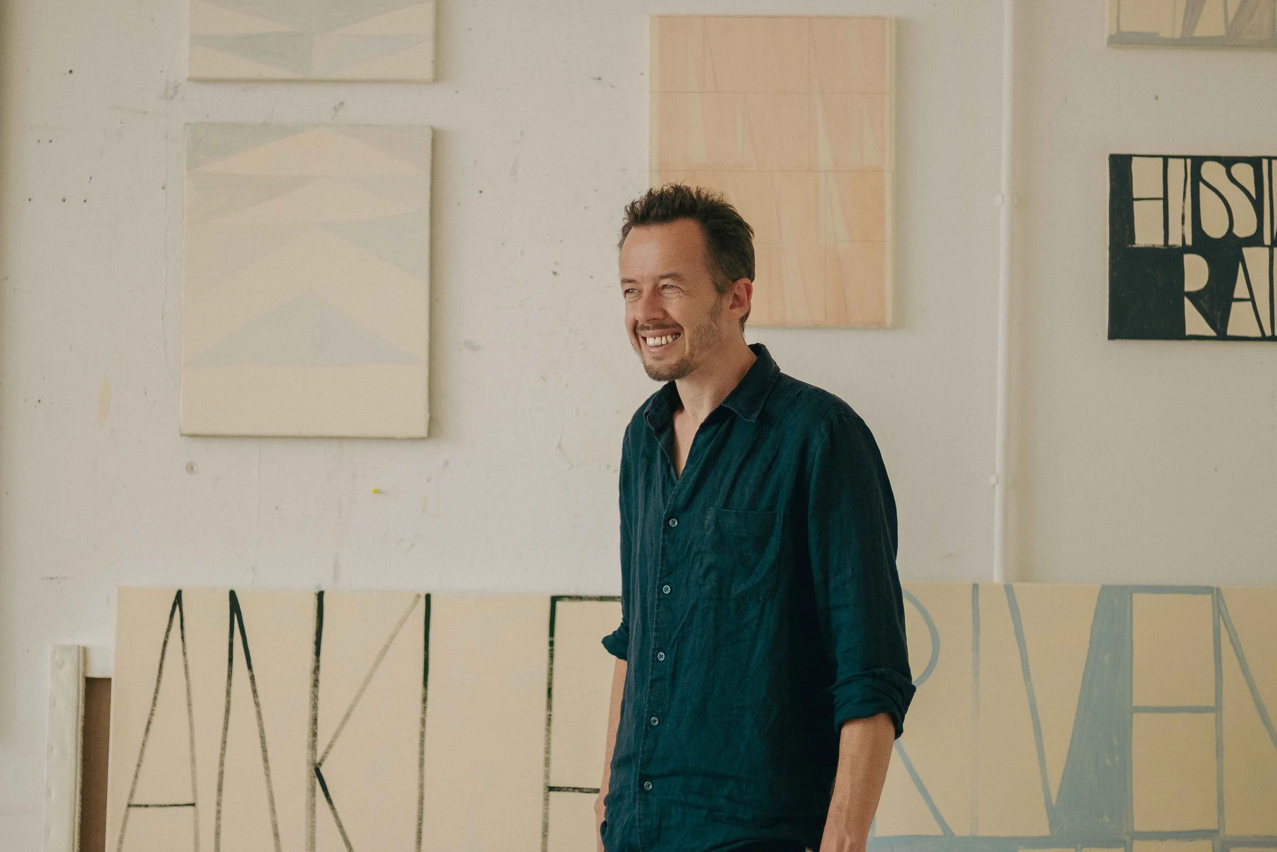 A smiling man with dark hair and a beard in a dark button-up shirt standing in front of a wall with abstract artwork.