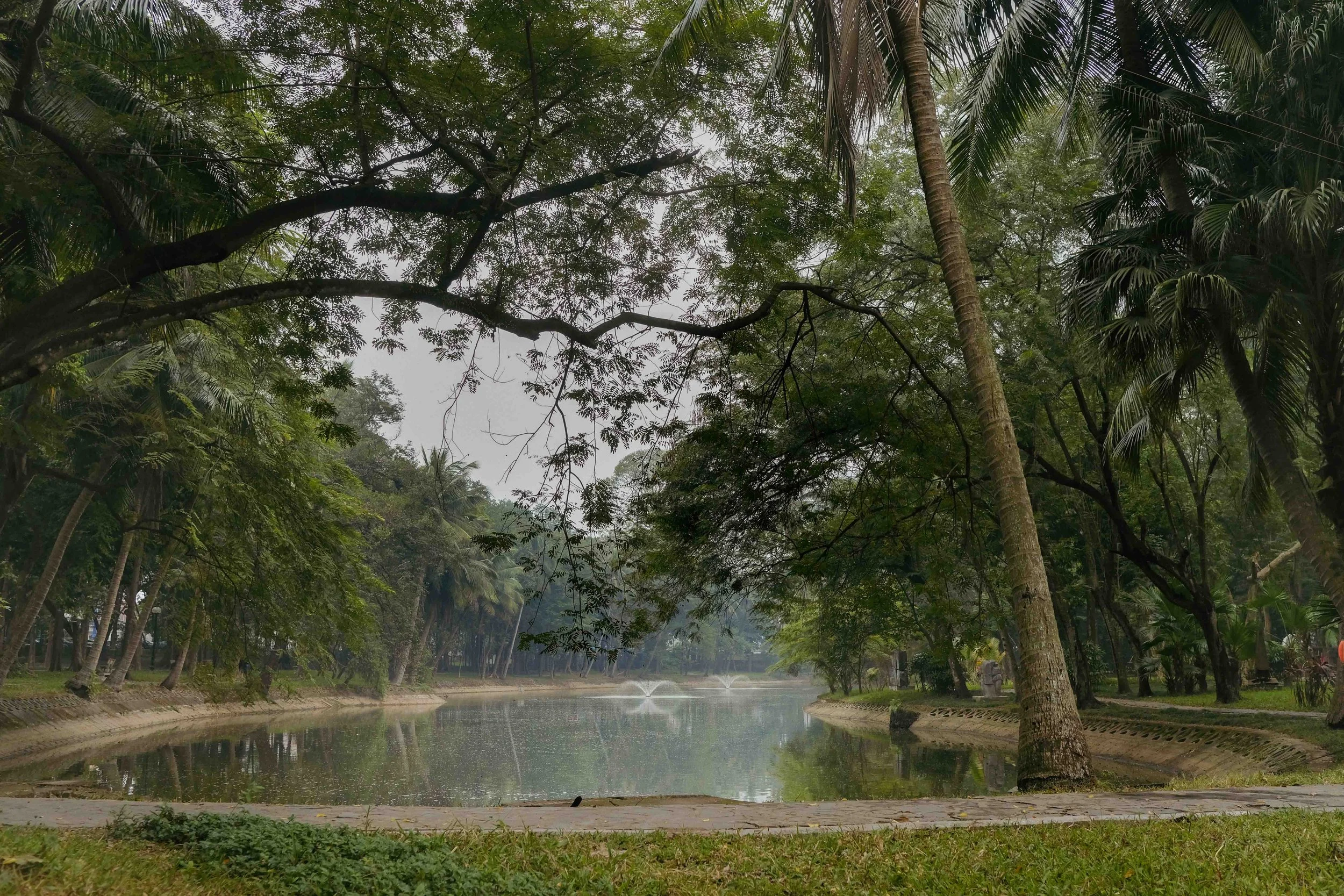 A peaceful park scene with a river, tall trees, and a fountain in the distance, under a cloudy sky.