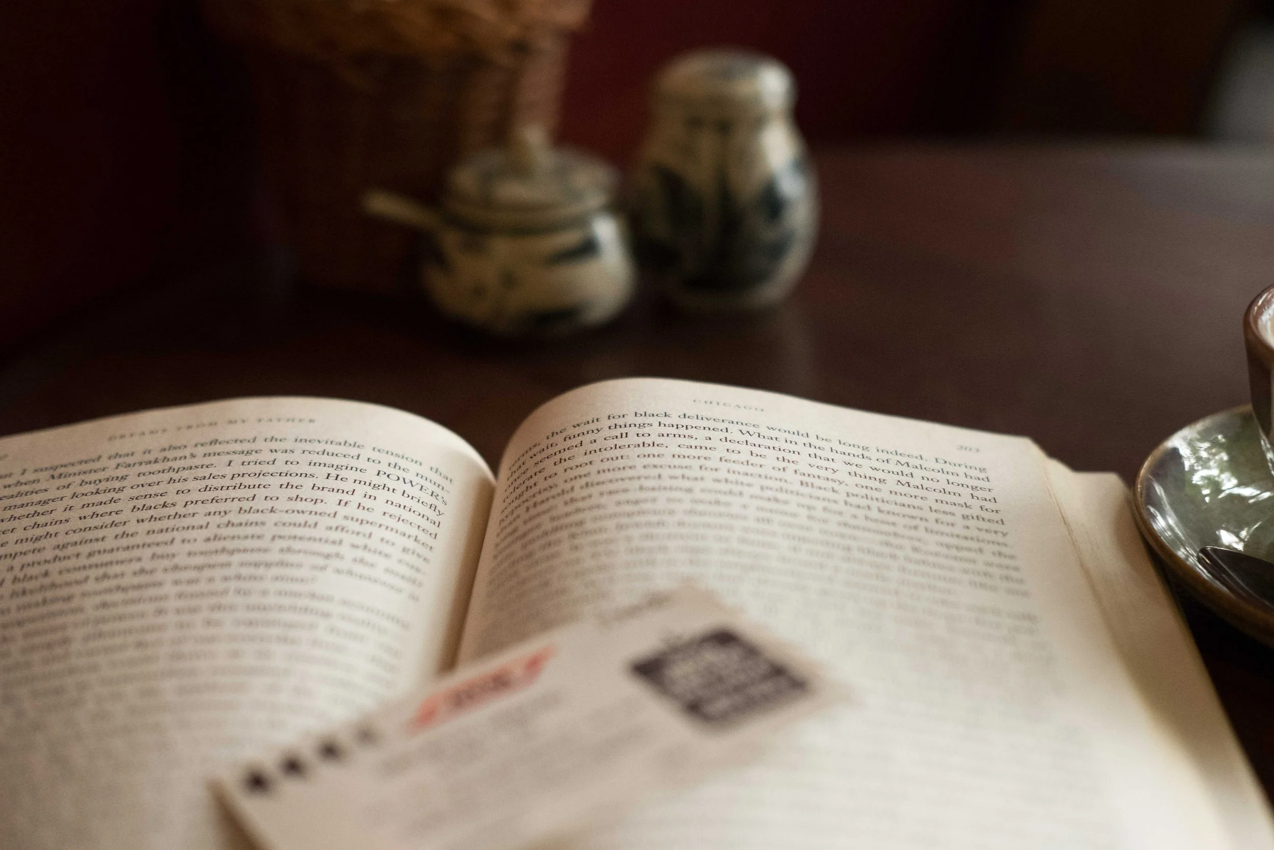 A close-up of an open book with a blurred background showing two small ceramic vases and a glass paperweight on a dark table.