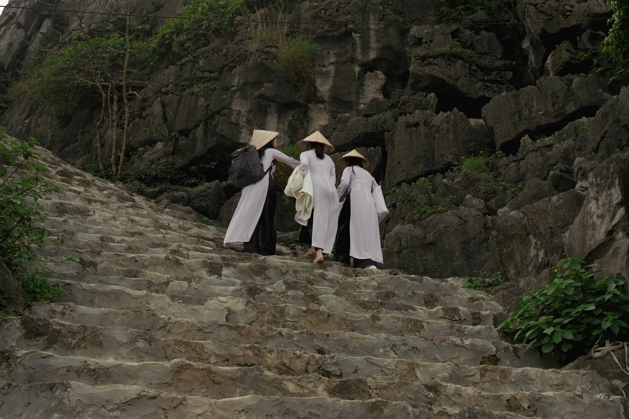 Four people wearing traditional conical hats and white clothing are climbing stone stairs in a rocky, forested area.