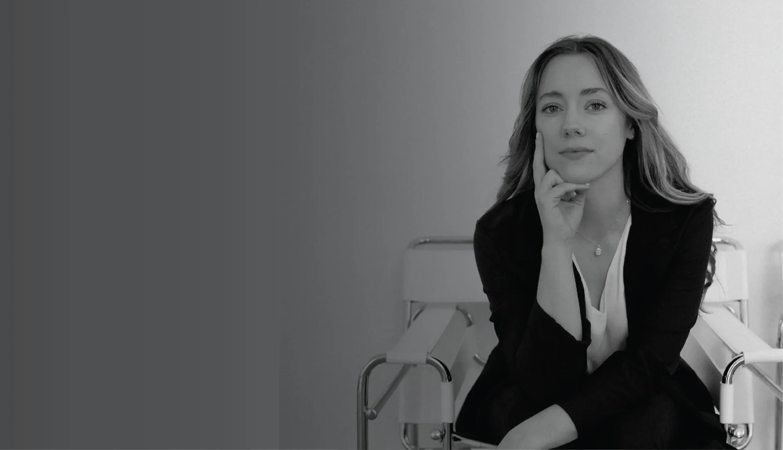Founder of WildHive Studio sitting on a white chair in a room, resting her chin on her hand and smiling slightly, with her other hand resting on her knee. She has long wavy hair and is dressed in business casual attire.