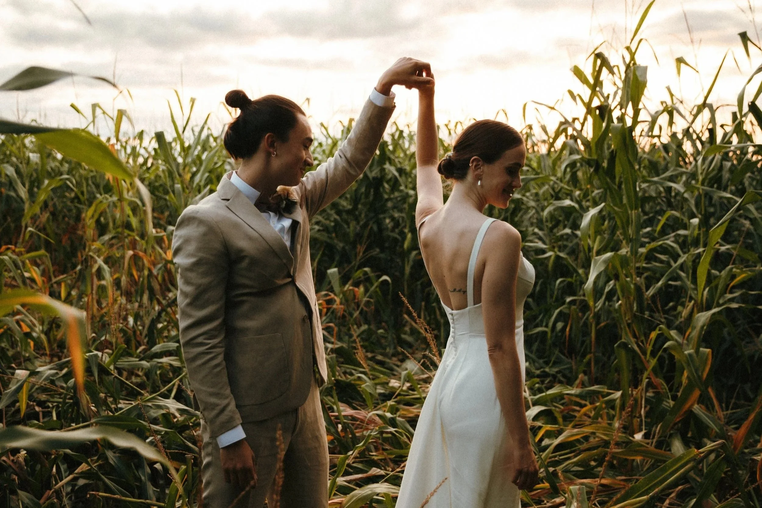 Deux personnes en tenue de mariage, un homme en costume beige avec un nœud papillon marron et une femme en robe de mariage blanche, se regardent tendrement dans un jardin.