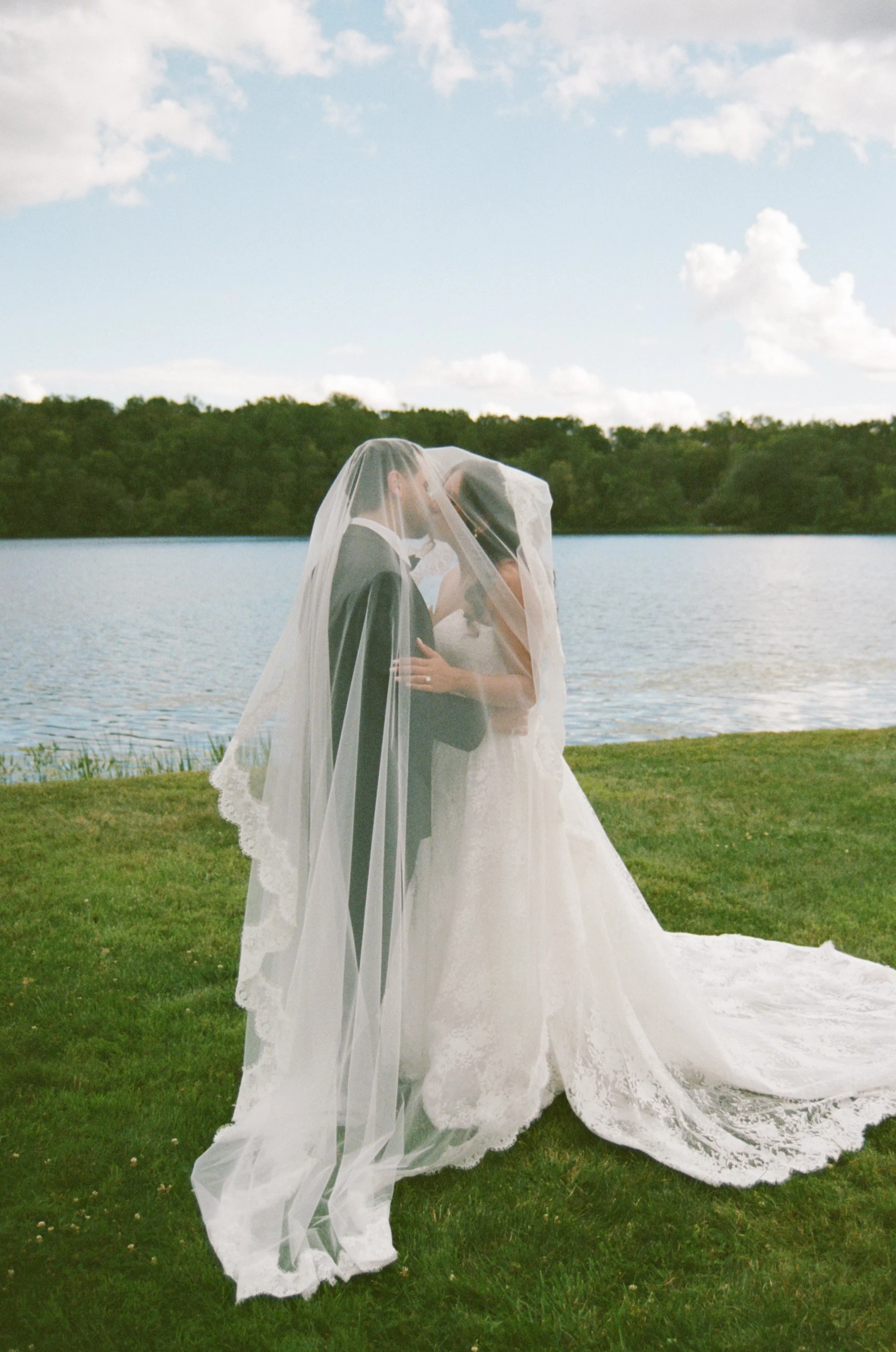 A bride and groom sharing a kiss outdoors near a lake, with the bride wearing a wedding gown and veil, and the groom in a tuxedo.