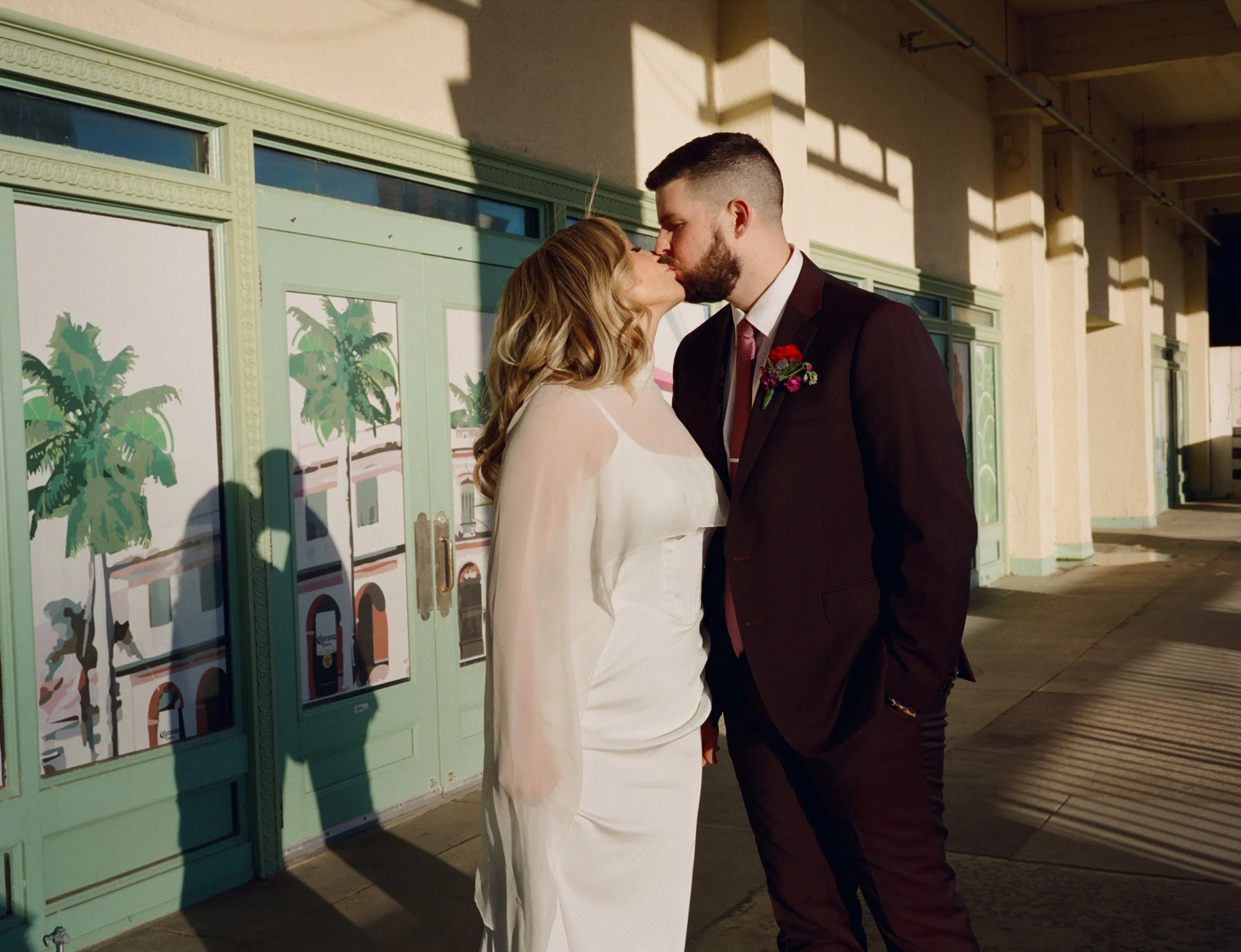 A couple shares a kiss outside a building with mint green window frames and palm tree illustrations on the glass, during late afternoon sunlight.