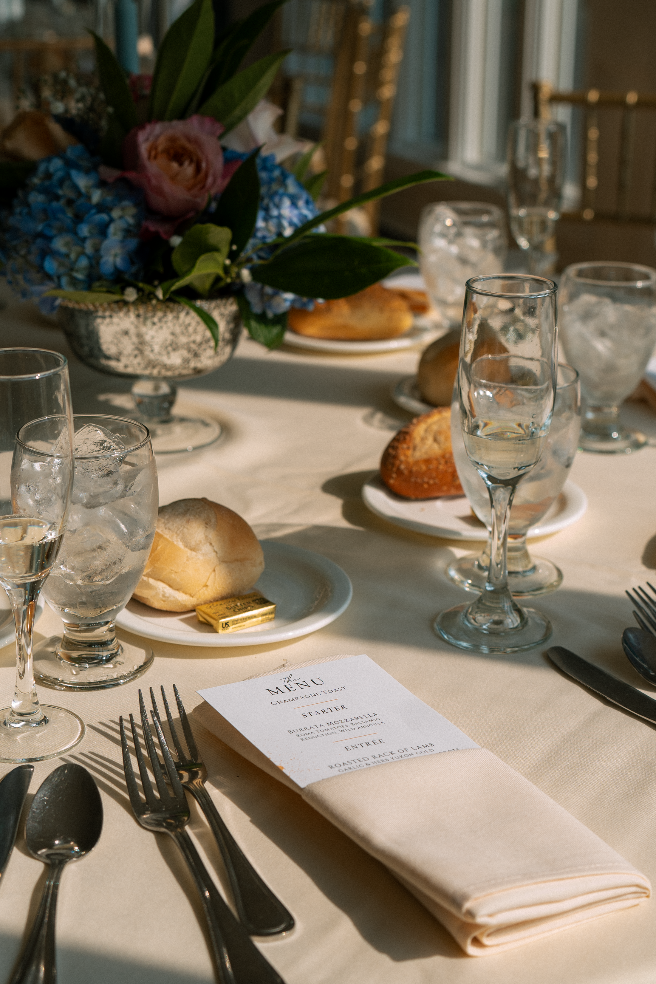 A close-up of a formal dining table setting with a white tablecloth, wine glasses filled with ice water, a floral centerpiece with pink and blue flowers, plates of bread, a gold-wrapped butter pat, and a printed menu. Silverware is arranged around th