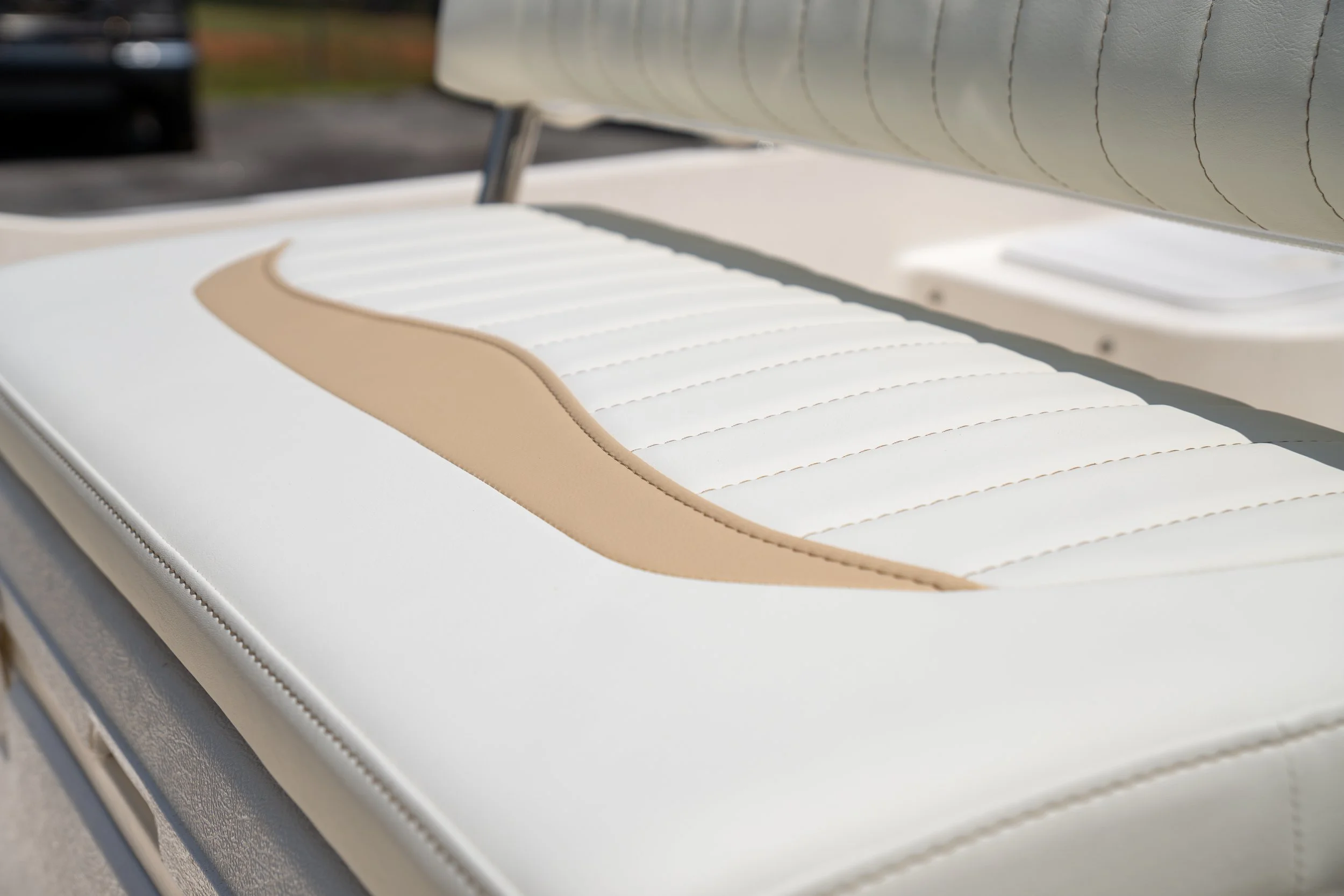 Close-up of a white leather bench seat with beige padding accents, likely on a boat or yacht, with a part of a gray metal surface and blurred background visible.