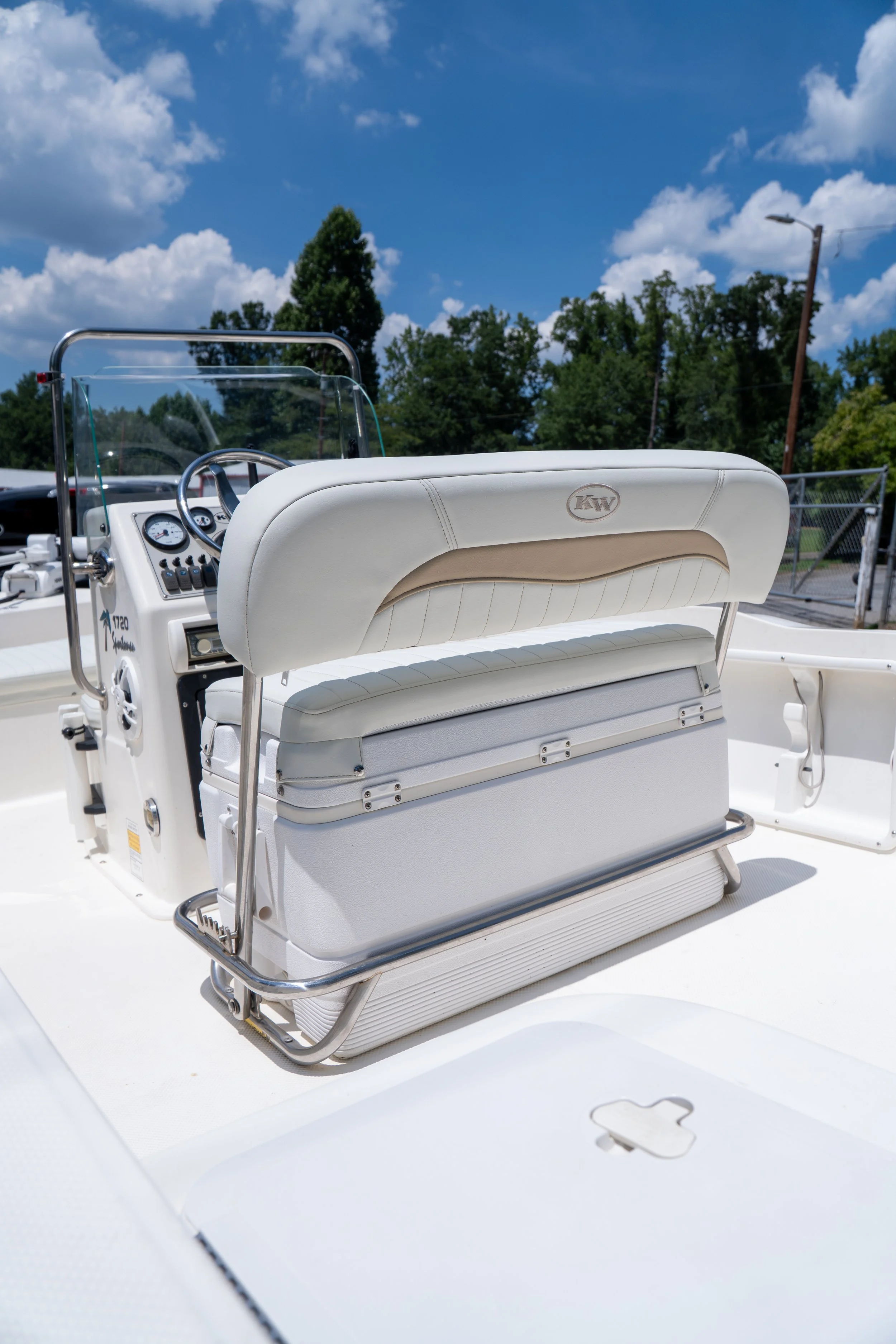 Close-up of a white boat dashboard with a title that says 'KW', steering wheel, gauges, and seating area. Outside, there are trees, a fence, and a clear blue sky with clouds.