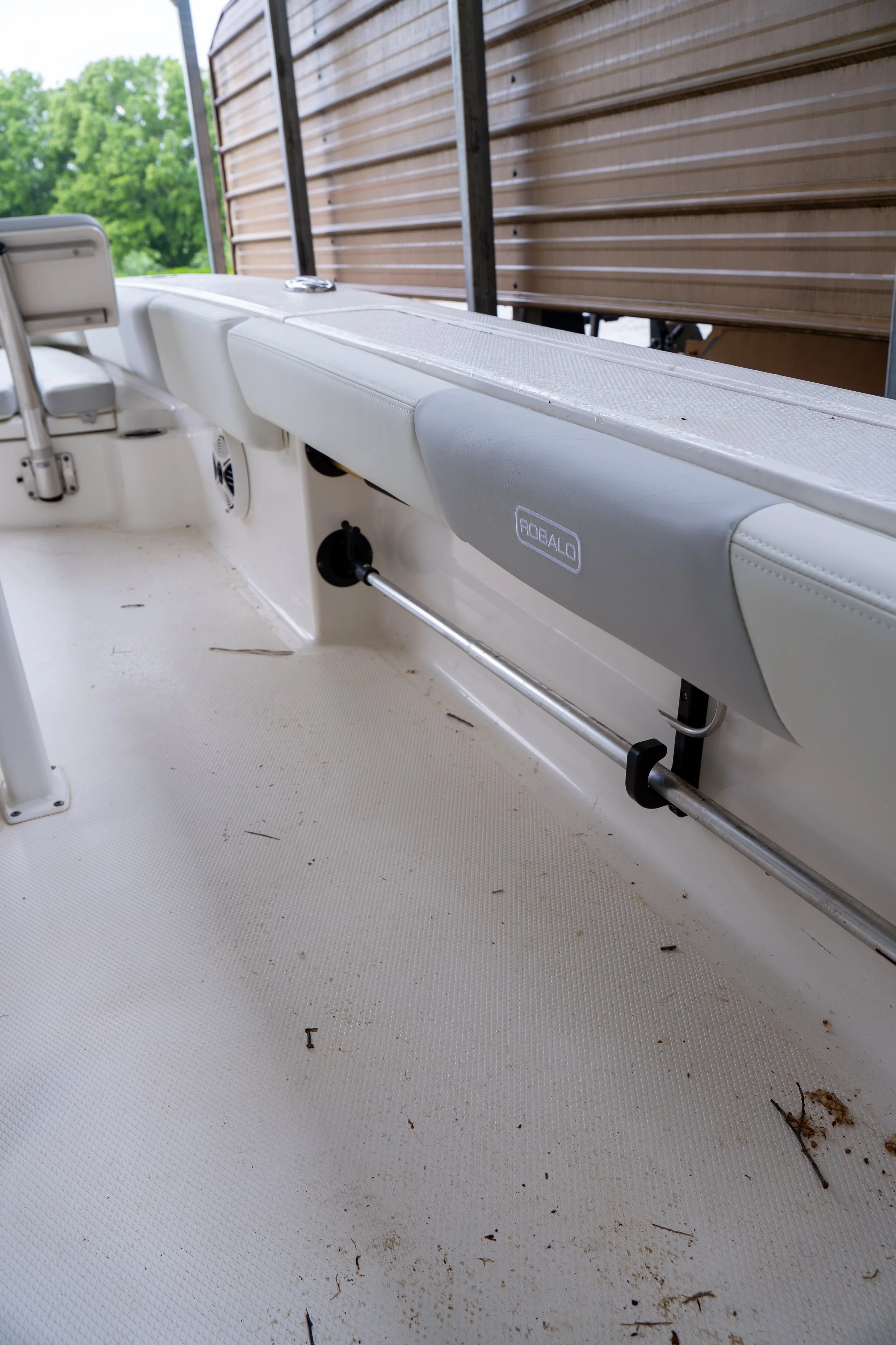 Interior of a boat with a white cushioned seat and a deck that has some debris, including small twigs and dirt, scattered on it. A brown wooden wall is visible in the background.