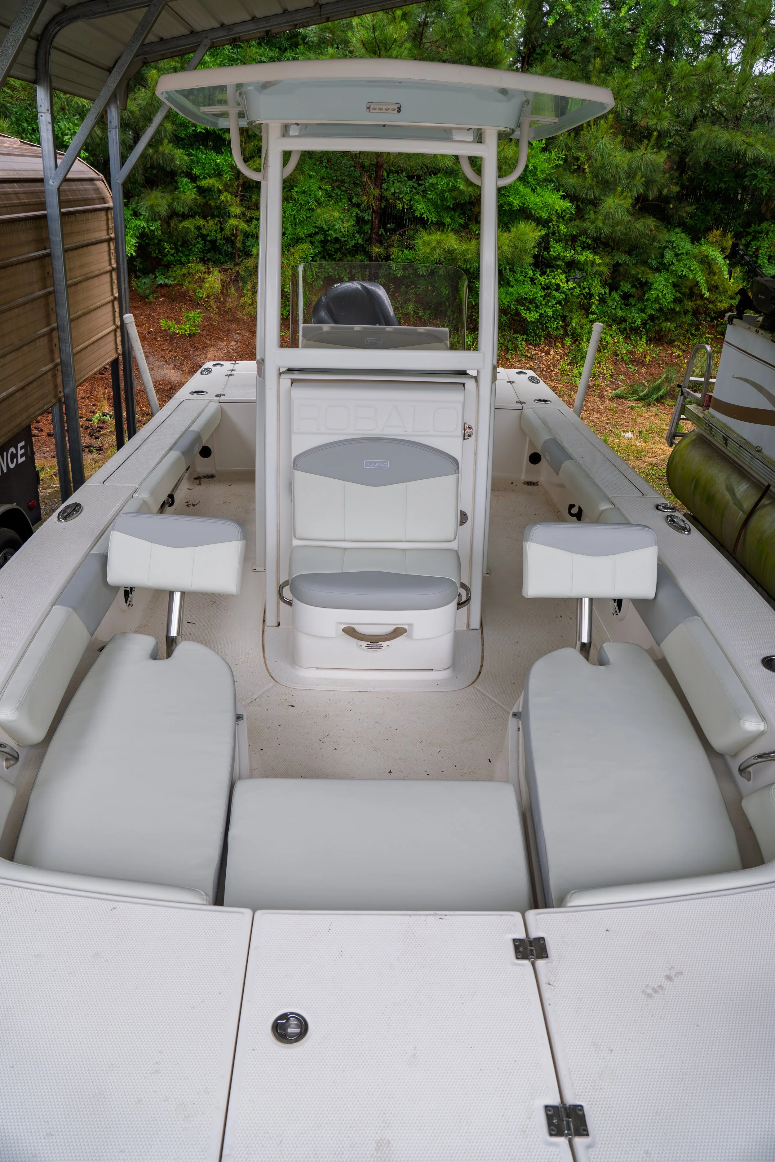White fishing boat with a center console, cushioned seats, and a T-top canopy, parked under a metal shelter with green trees in the background.
