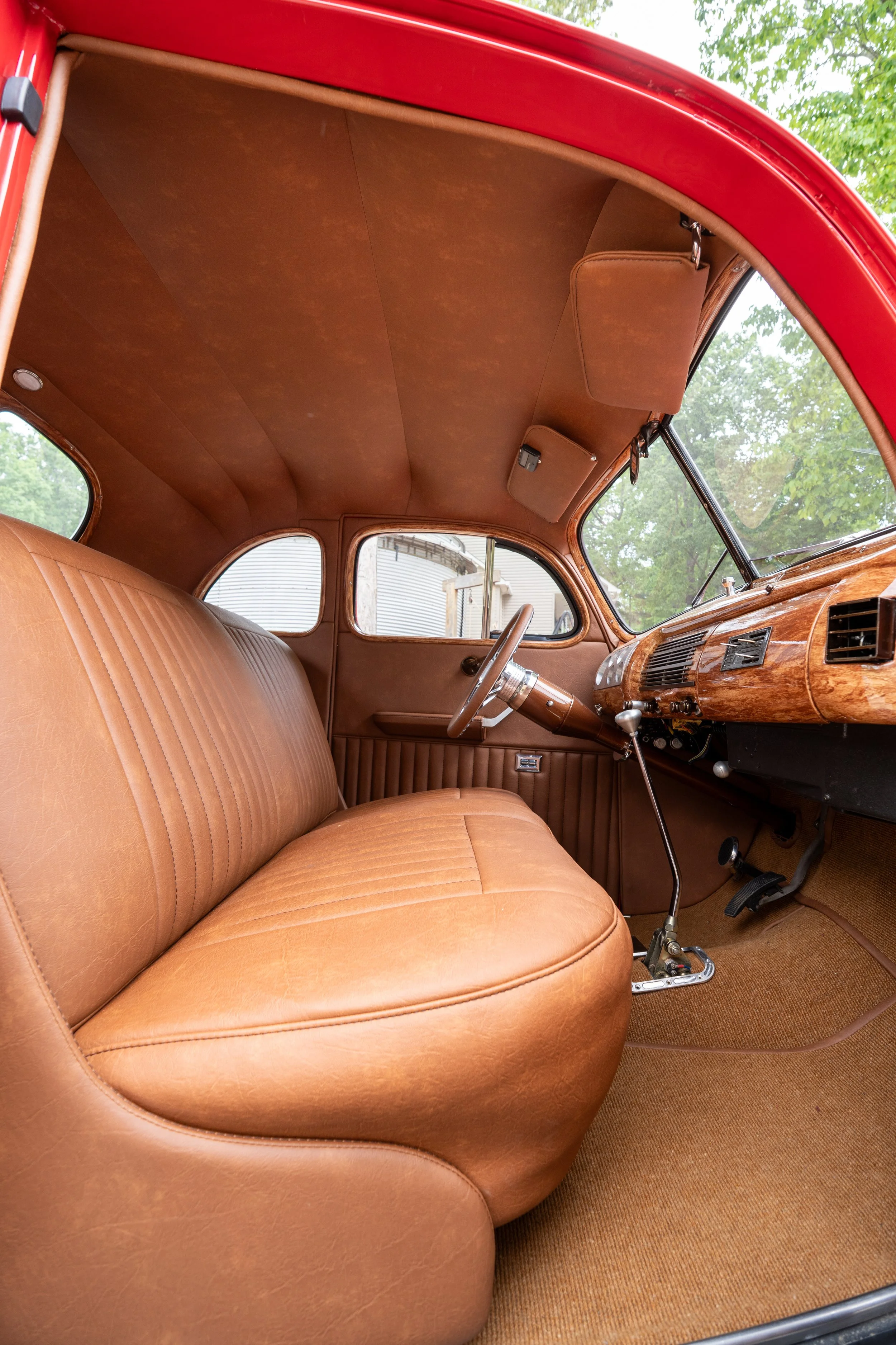 Interior of a vintage car with tan leather seats, wooden dashboard, and brown fabric ceiling, viewed from the passenger side looking toward the driver.