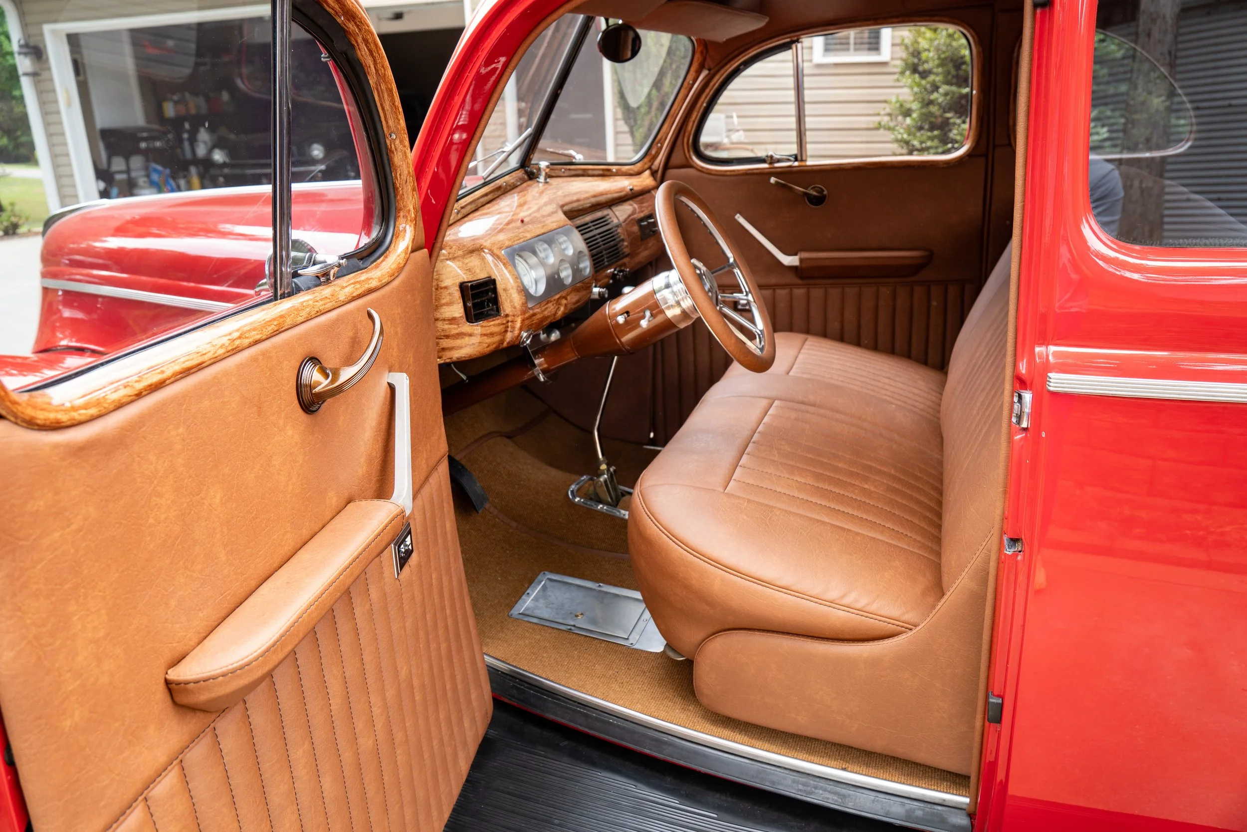 The interior of a vintage red car with tan seats, wooden dashboard, and steering wheel, parked outdoors next to a house.
