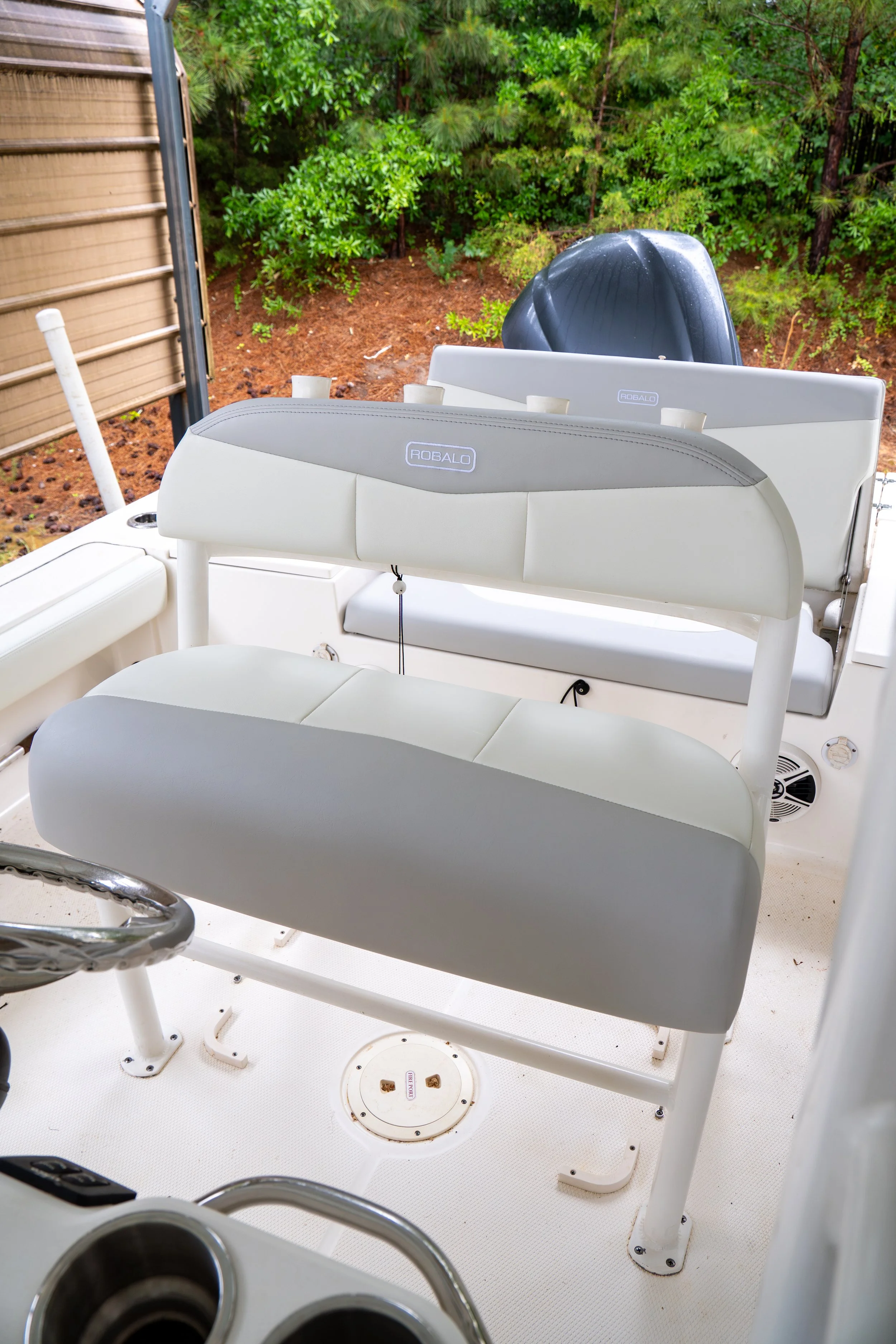 The image shows the interior deck of a boat with two white cushioned seats and a black outboard motor at the stern, surrounded by green trees and shrubs.