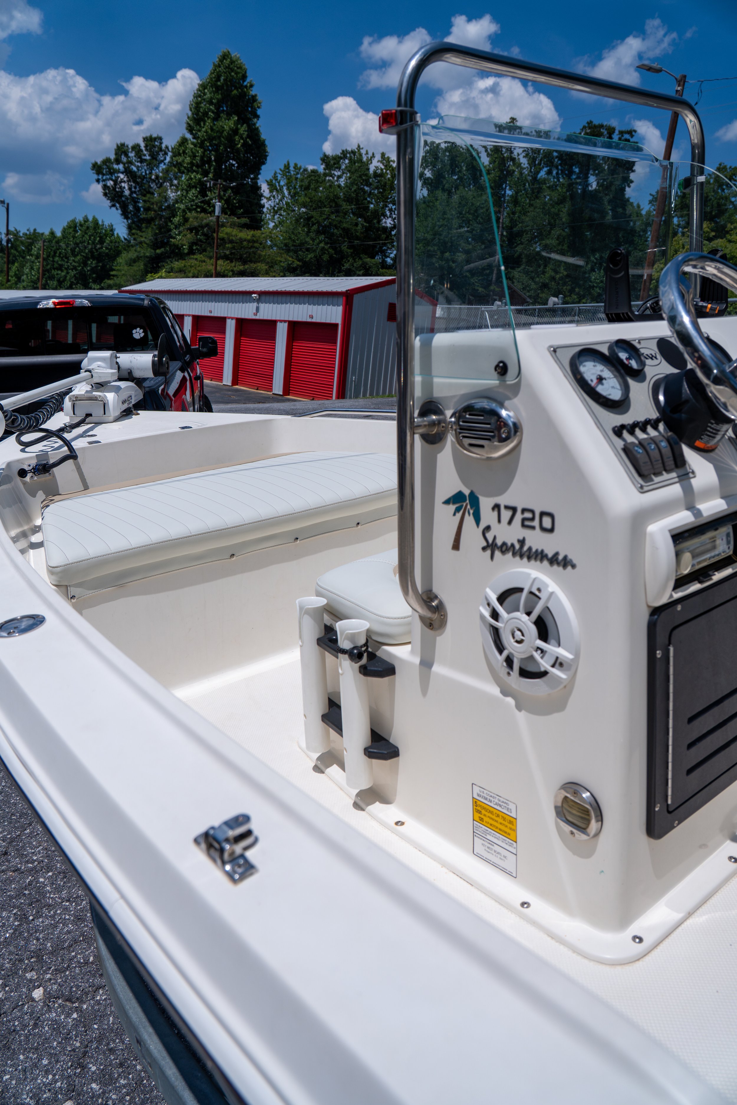 Close-up of a small white boat's control panel with gauges and switches, and a partially visible red and white storage building in the background under a sunny blue sky with clouds.