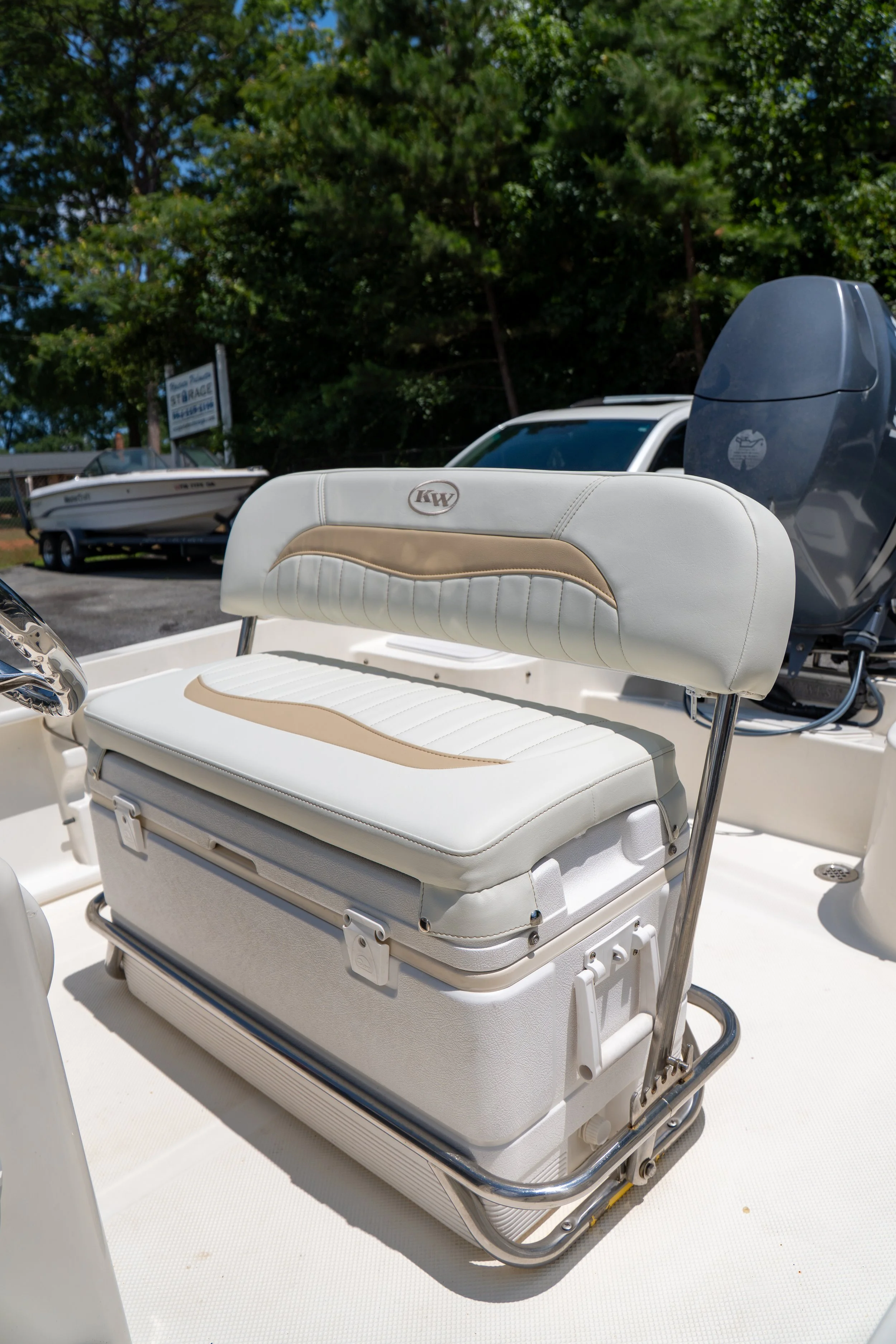 A white boat with a cushioned seat in the foreground, and a boat motor and vehicle parked in the background under trees.