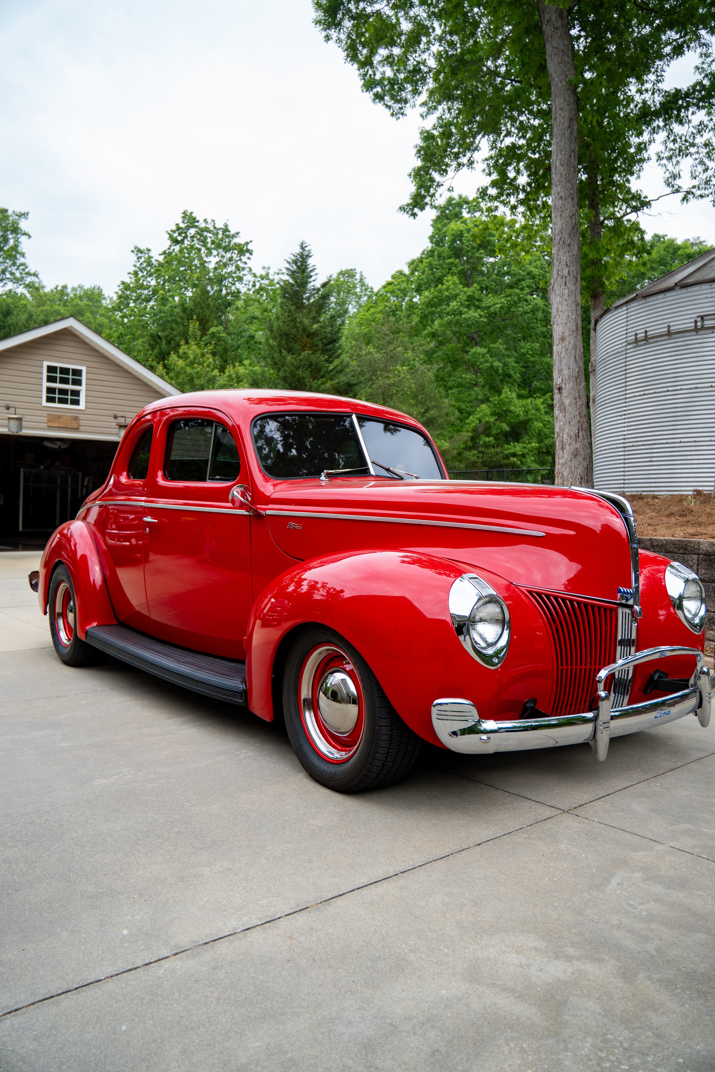 A bright red vintage Ford automobile parked on a driveway with a house and green trees in the background.