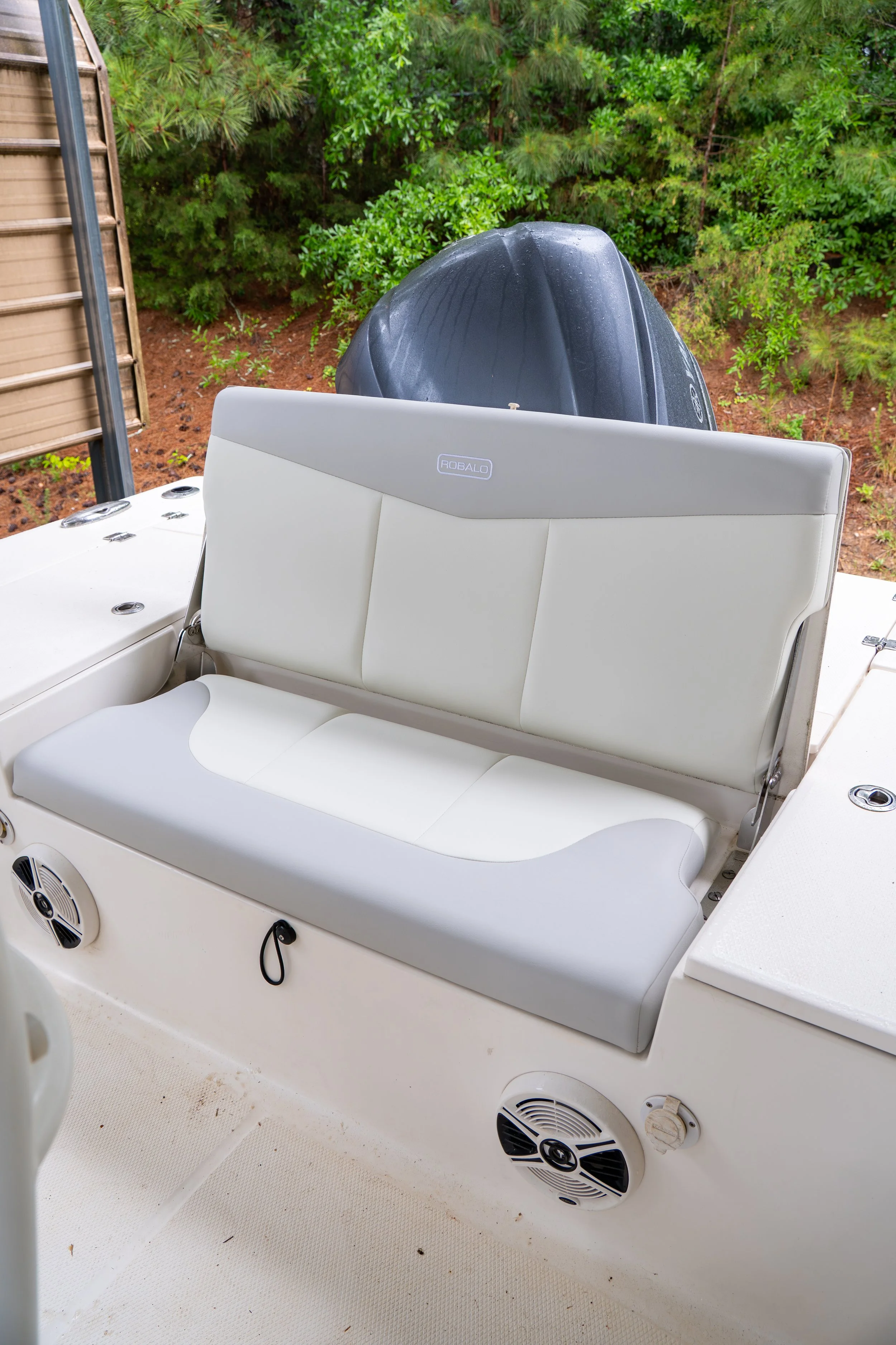 The image shows the back of a boat with a white cushioned seat, a black outboard motor, and a background of green trees and bushes. The boat's interior has built-in speakers and some small dirt particles on the floor.
