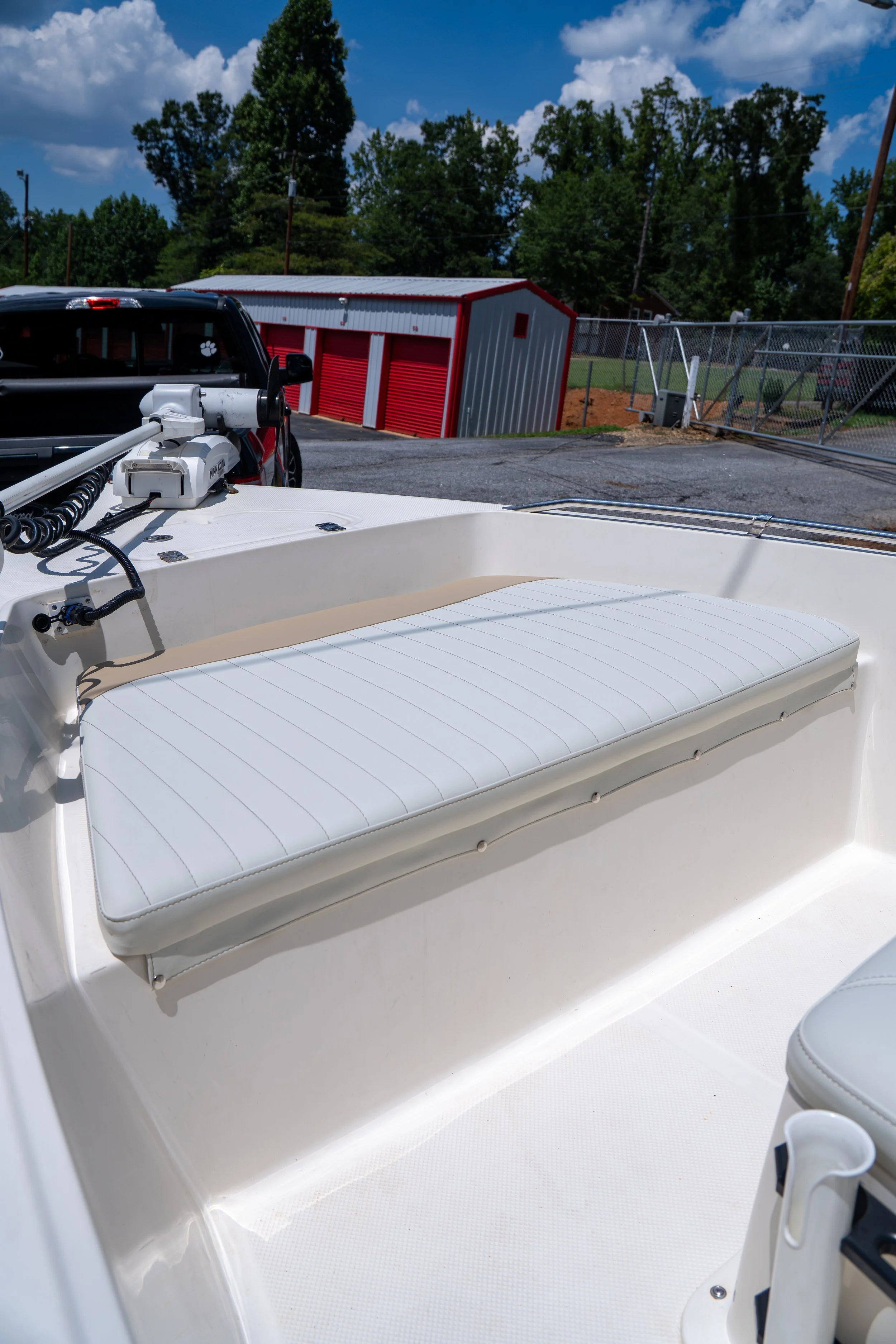 View of the interior of a boat showing a white cushioned seat, a mounted equipment on the boat's surface, and a view of a parking lot and a red and white shed with trees and blue sky in the background.
