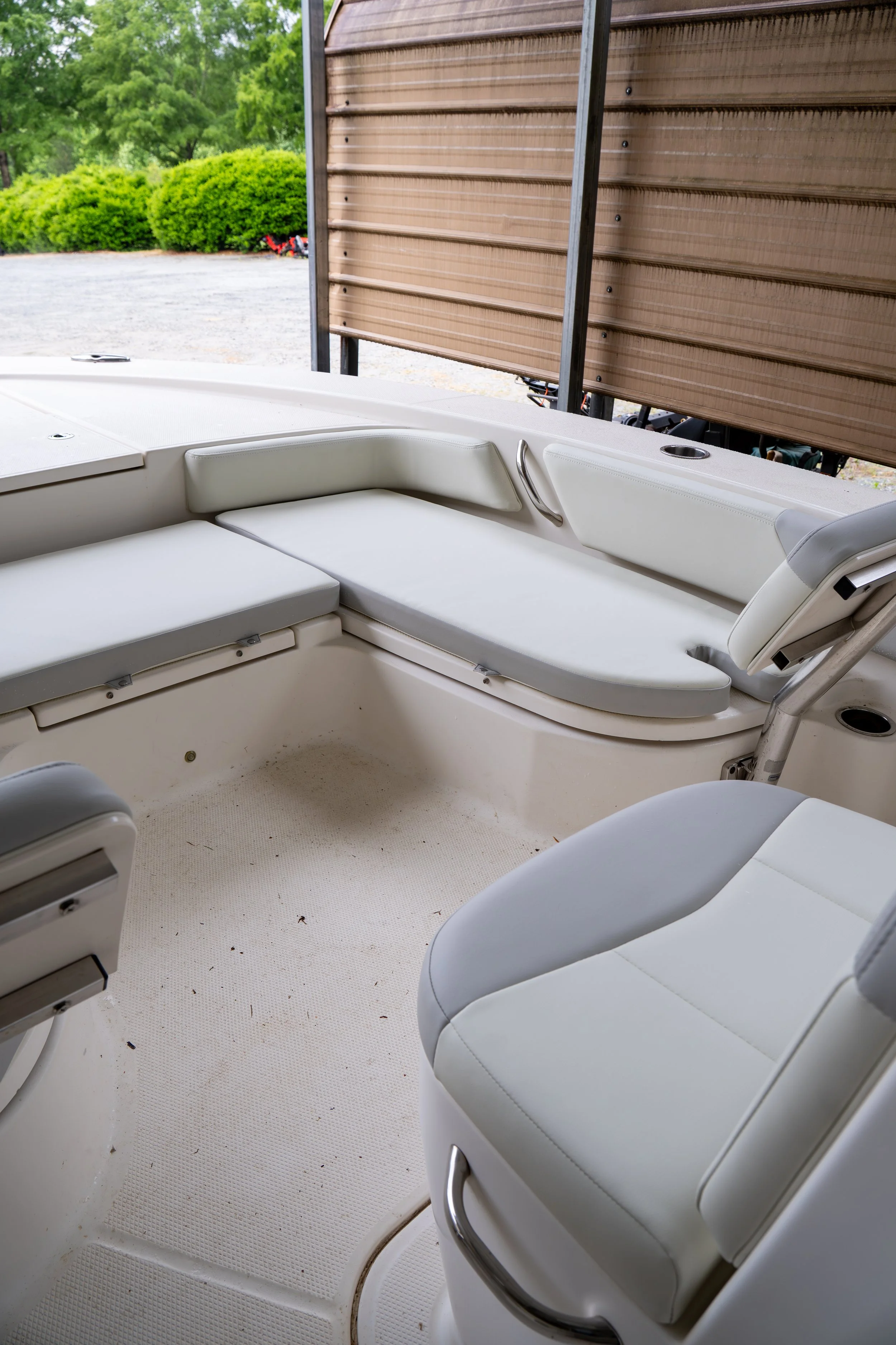 Interior view of a boat with white cushioned seats, metal handrails, and a textured white floor. Outside, there are green trees and shrubbery, and part of a wooden structure or wall is visible.